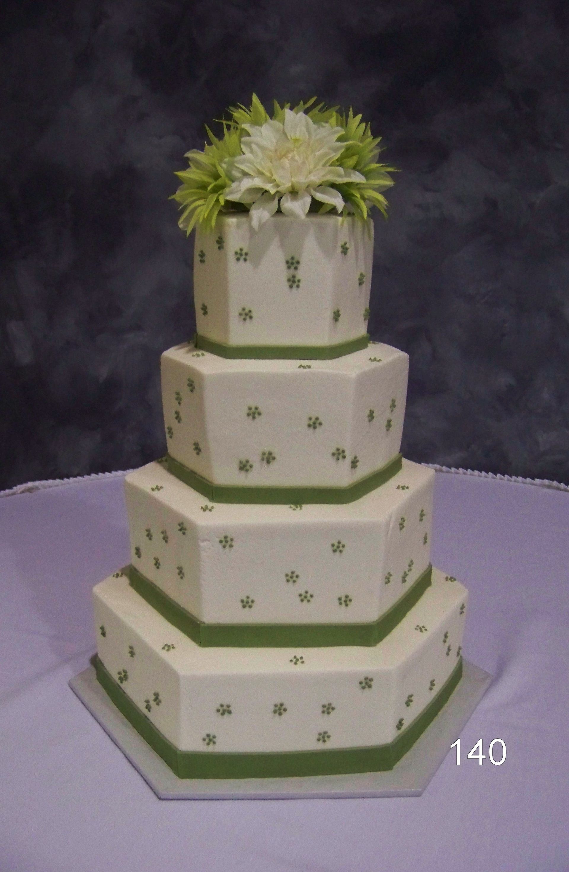 White tiered cake with green trim and small flower accents, topped with white and green flowers on a lavender tablecloth