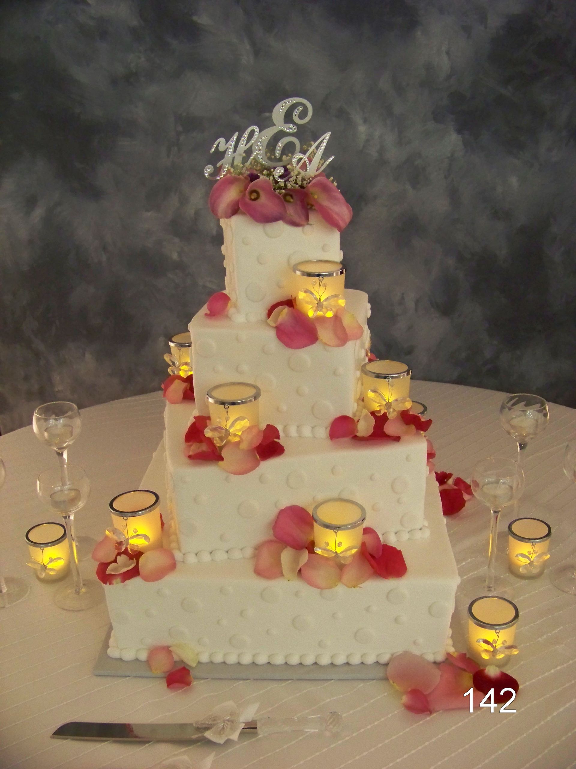 Three-tier white cake with pink flowers and lit candles on a white table, against a dark backdrop