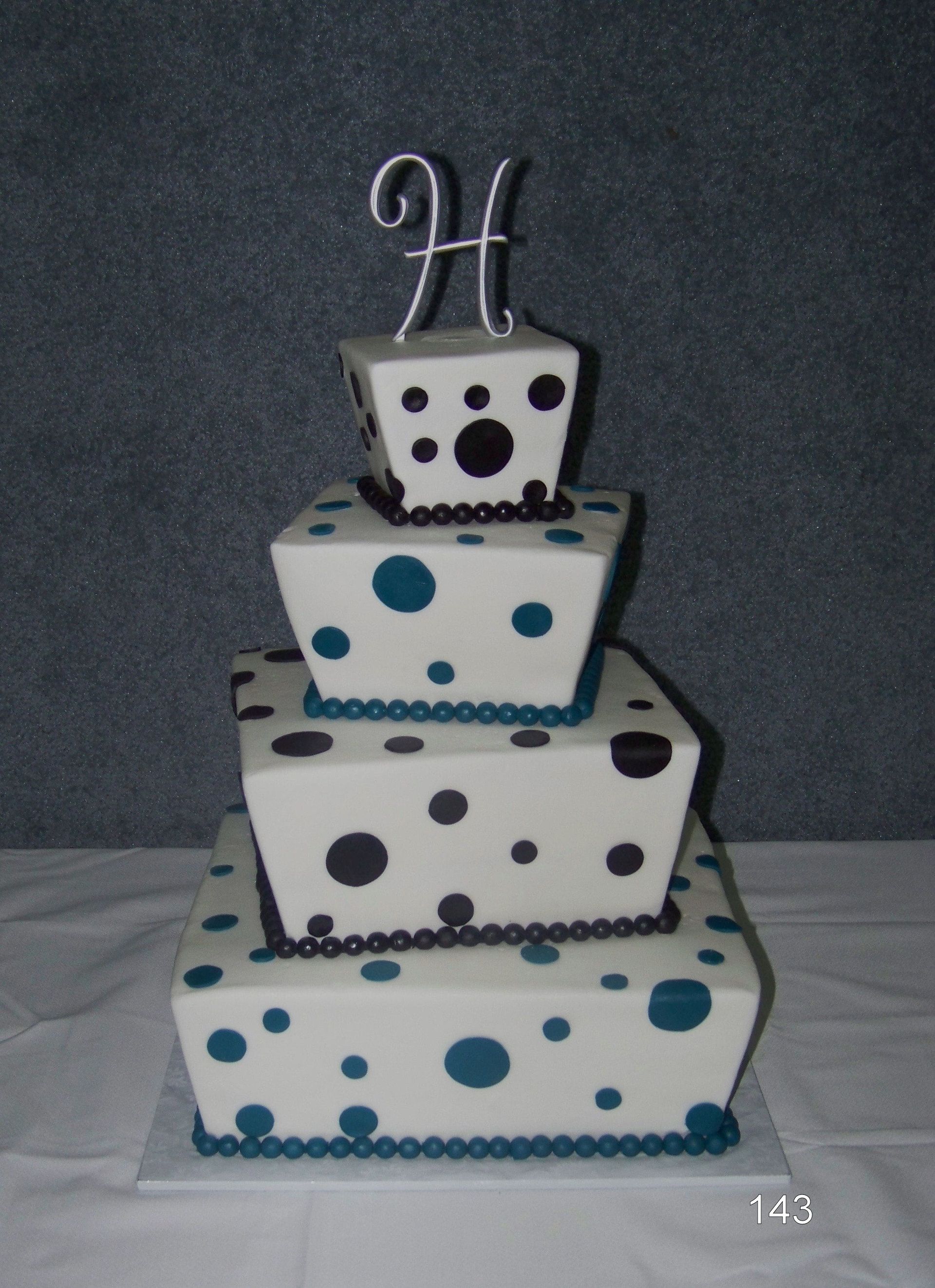 Tiered white cake with blue and black polka dots and a decorative silver topper on top.