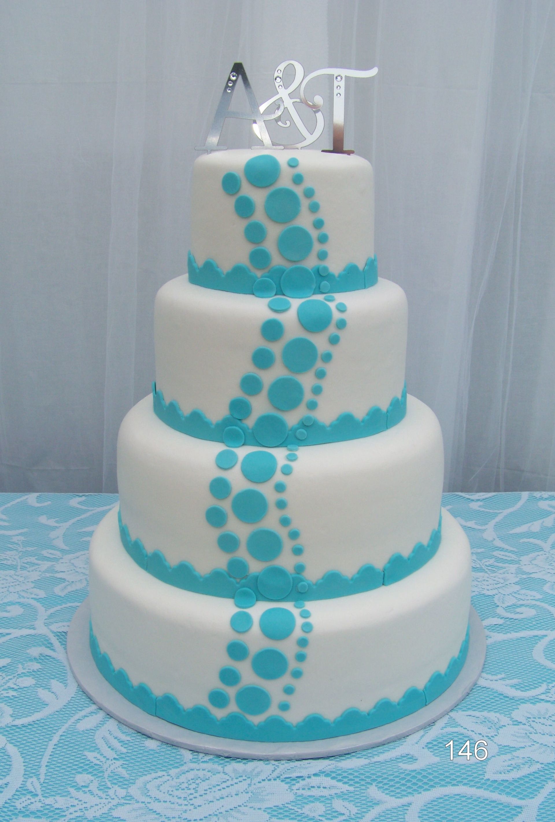Four-tier white cake with turquoise polka dots and silver topper on a table.