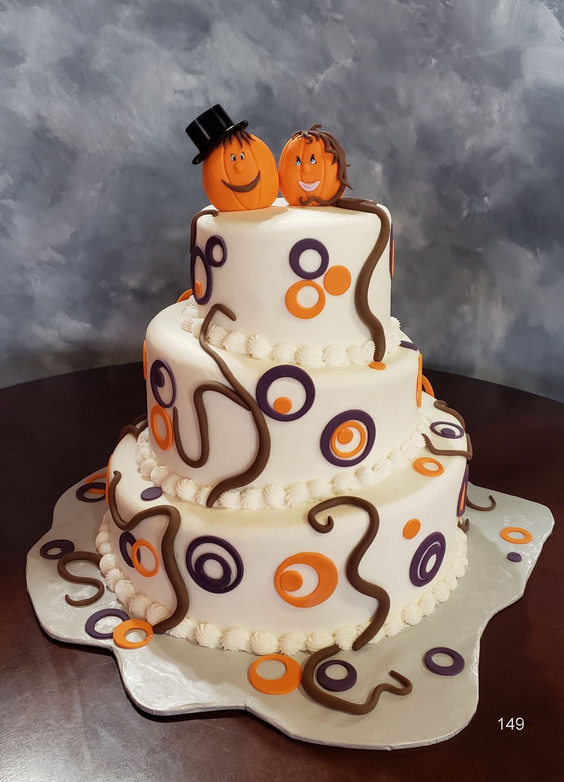 White tiered cake with orange and purple swirls, topped with two smiling pumpkins and a tiny black hat.