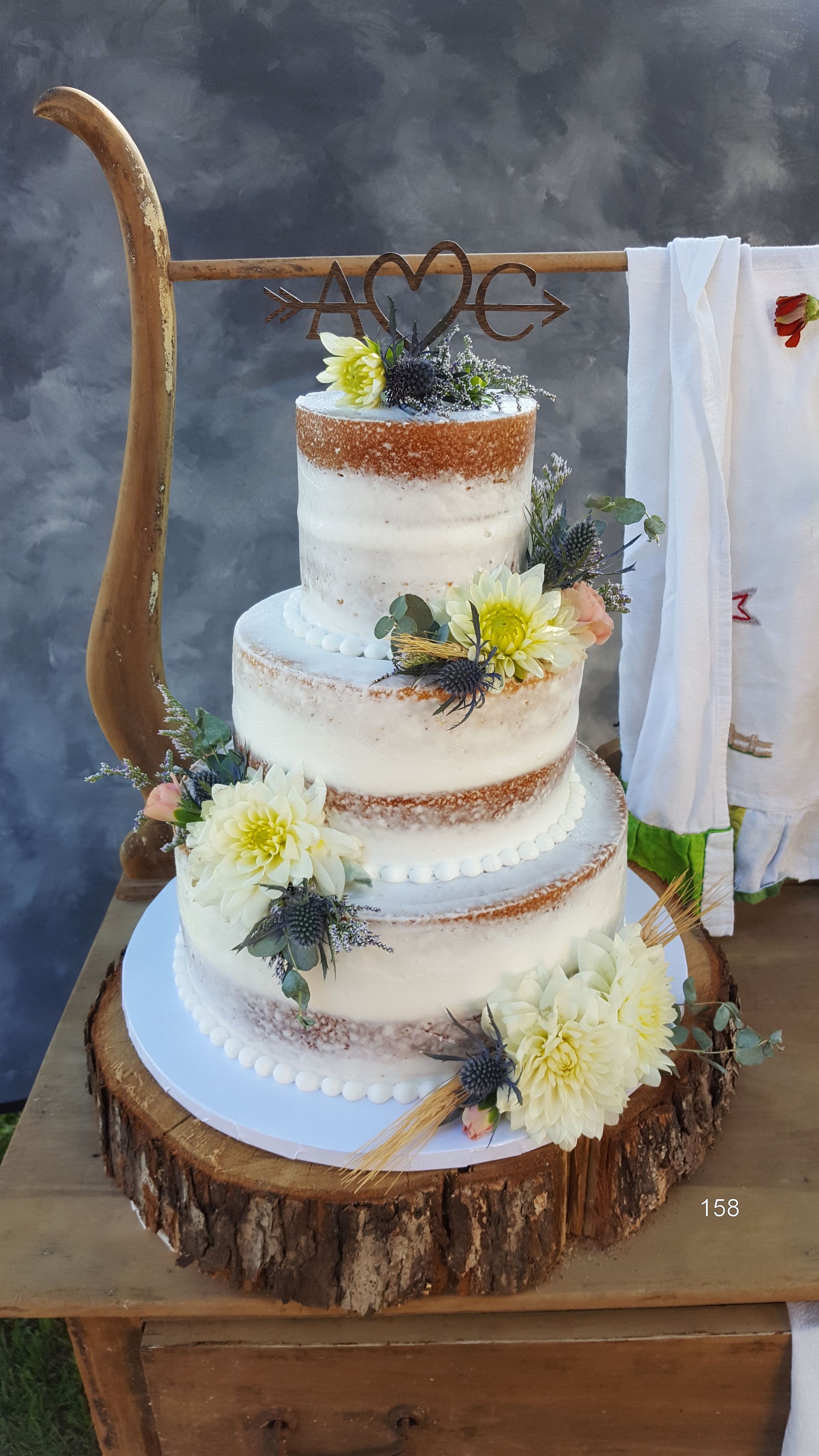 Three-tier white wedding cake on rustic wood, decorated with yellow flowers and a heart topper.