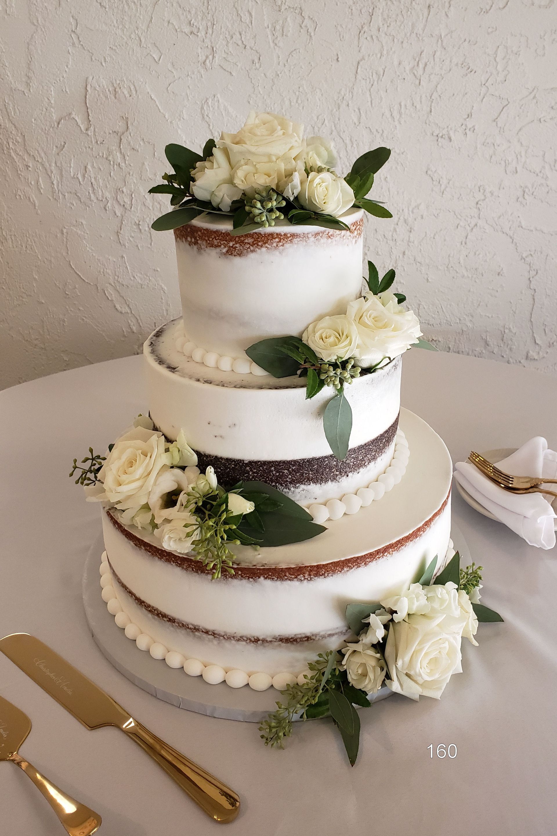 Four-tier white wedding cake with cream flowers and greenery on a table with gold utensils