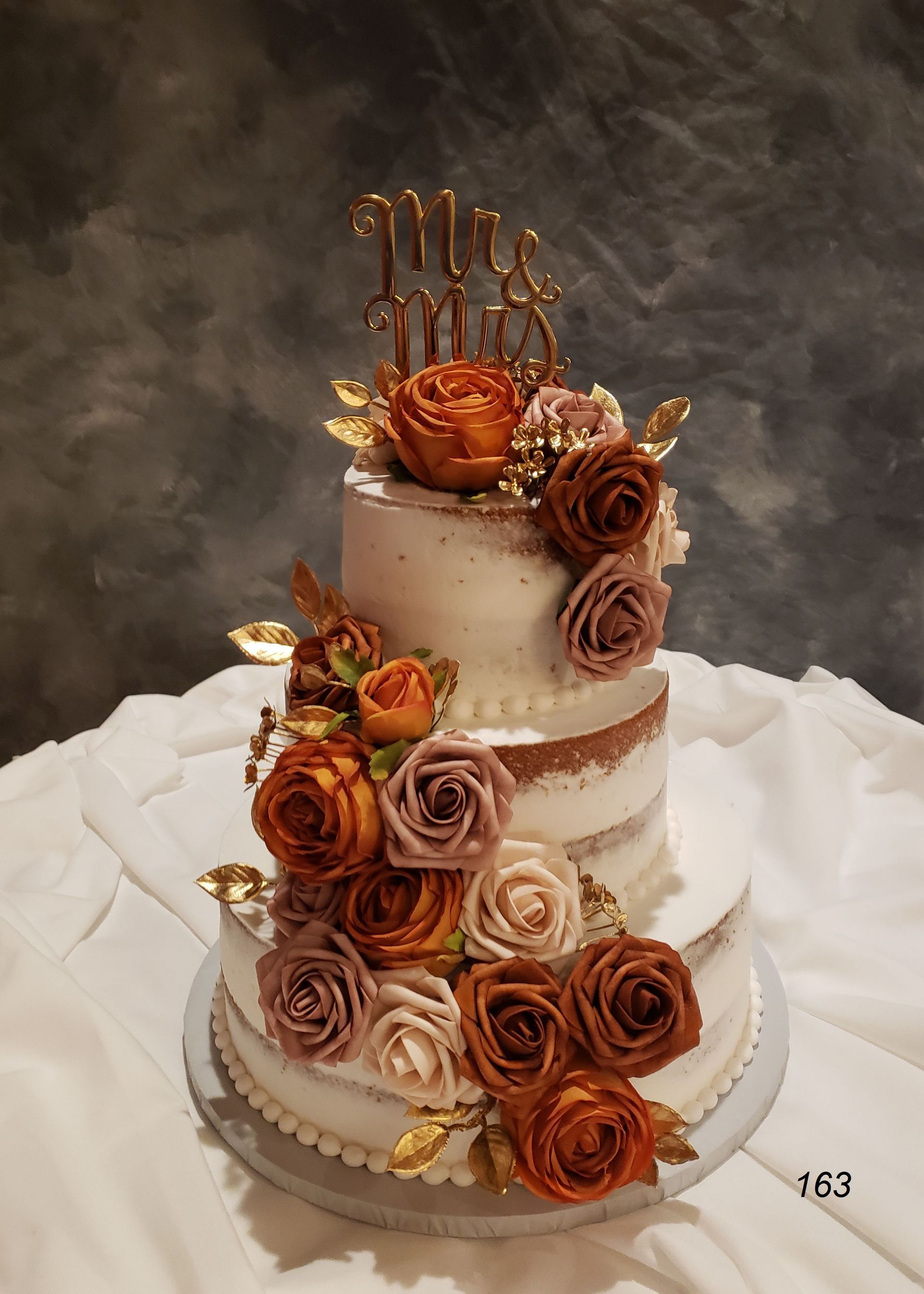 Three-tier white cake with copper rose decorations and a gold “Miss” topper on a white draped table