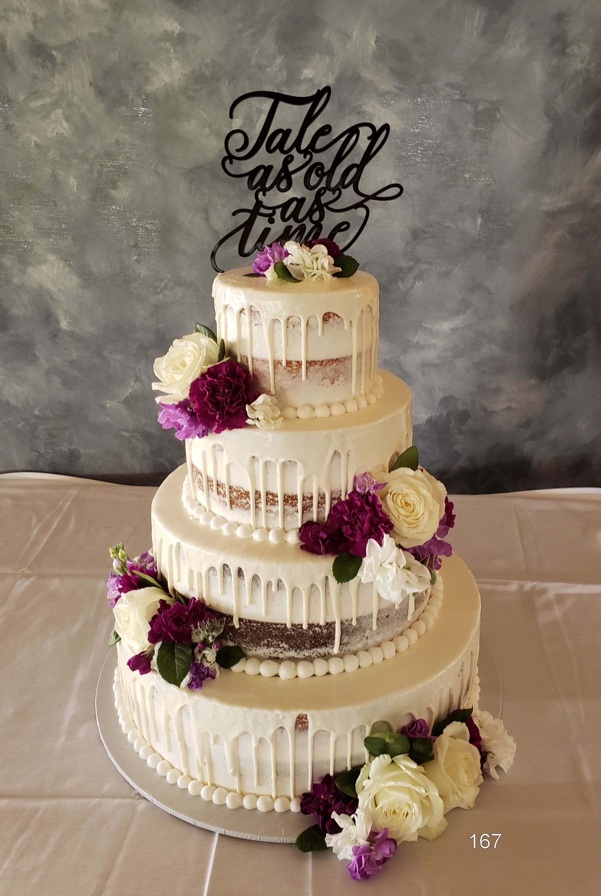 Four-tier white wedding cake with purple and cream flowers and black calligraphy topper