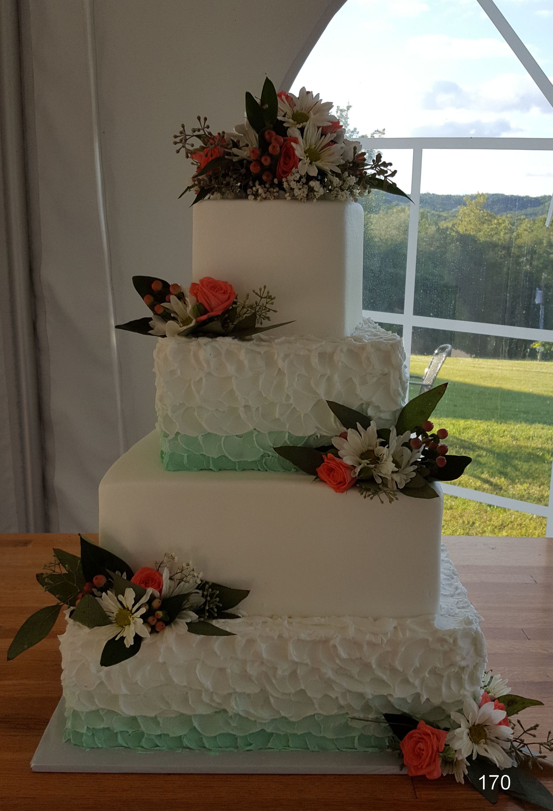 Four-tier white wedding cake with floral decorations and green ribbon trim by a bright window