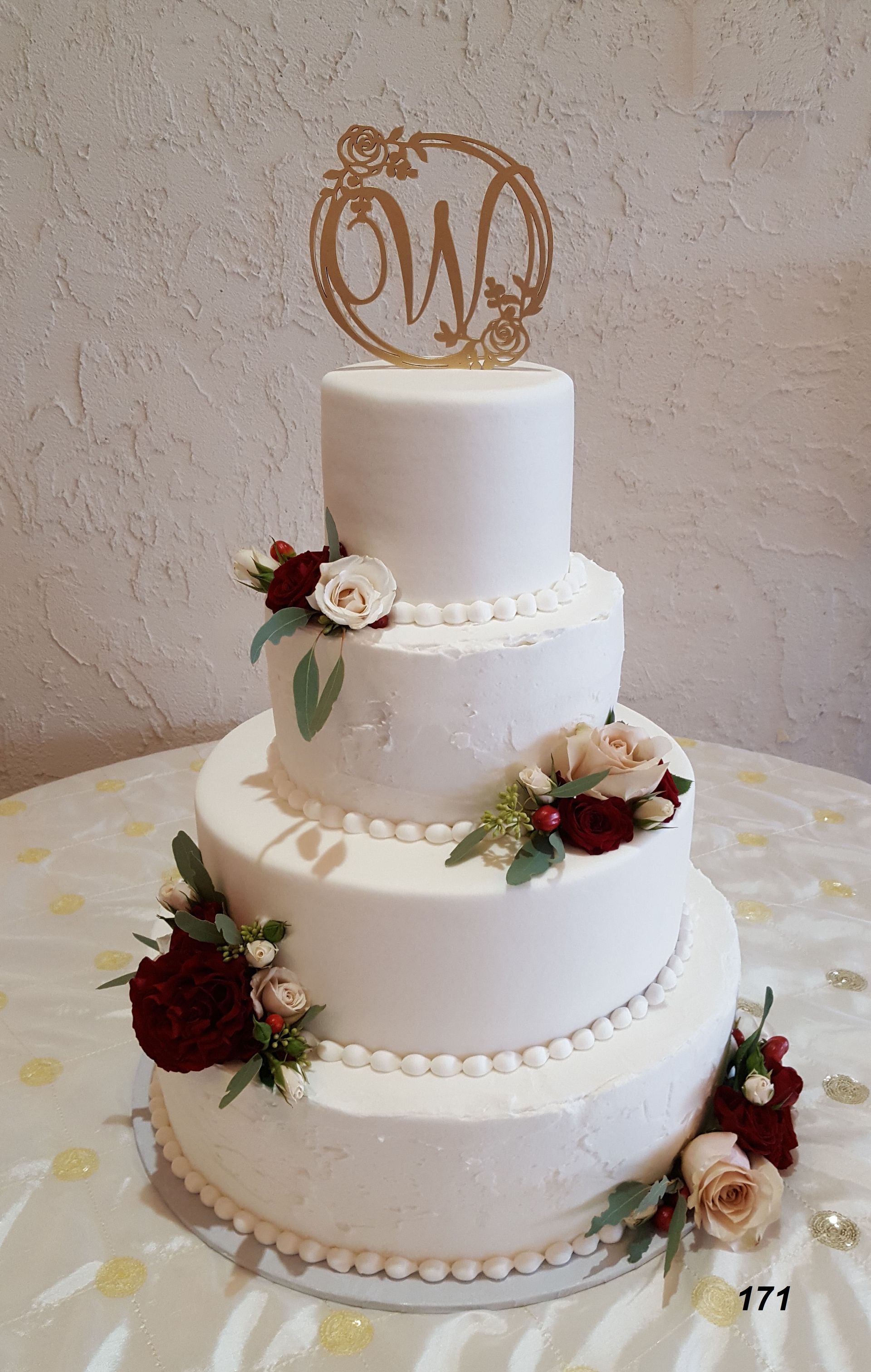 Three-tier white wedding cake with red and white flowers and gold monogram topper
