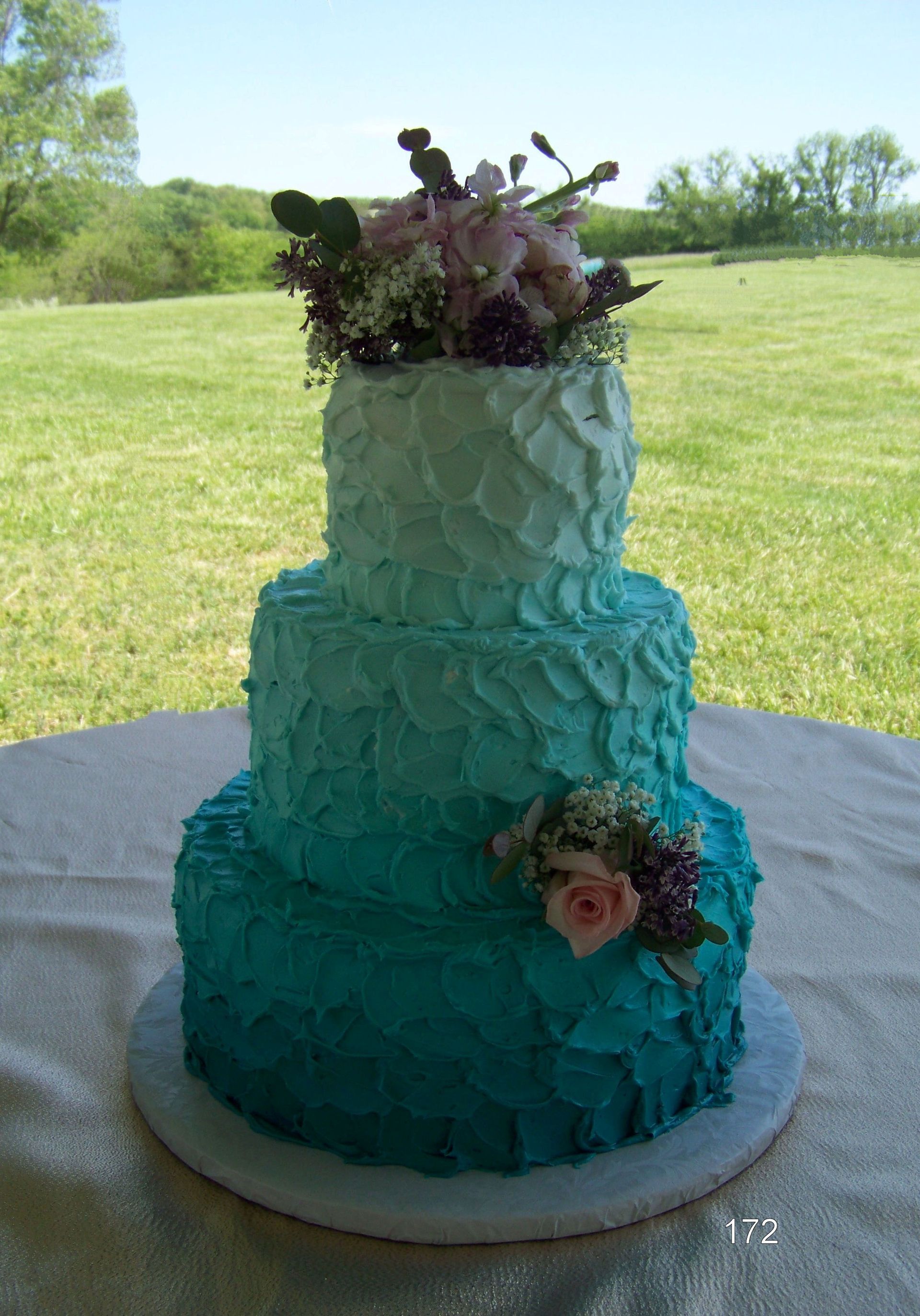 Three-tier turquoise wedding cake with purple flowers on a table outdoors