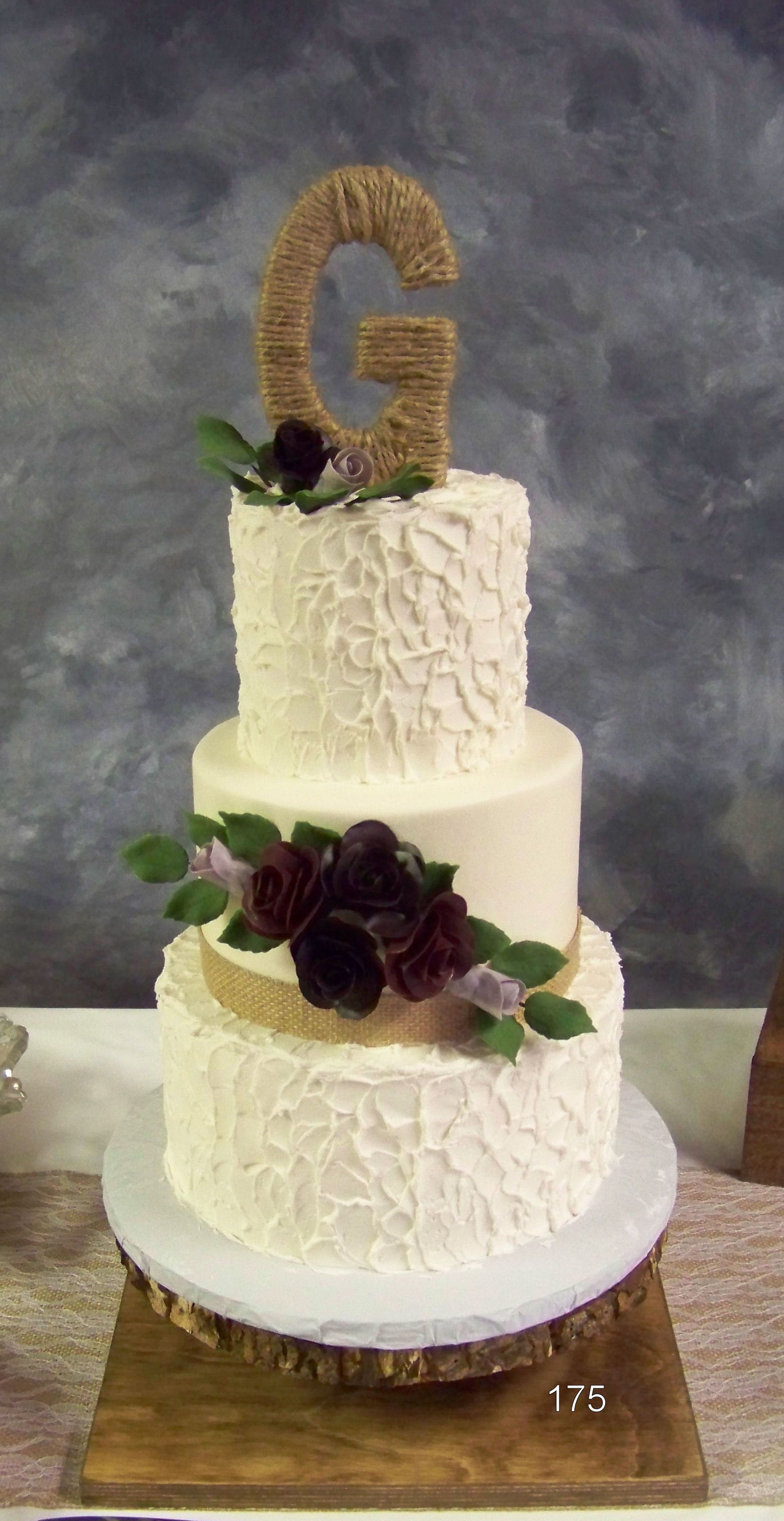 Two-tier white cake with gold letter G topper and dark floral decorations on a wooden stand
