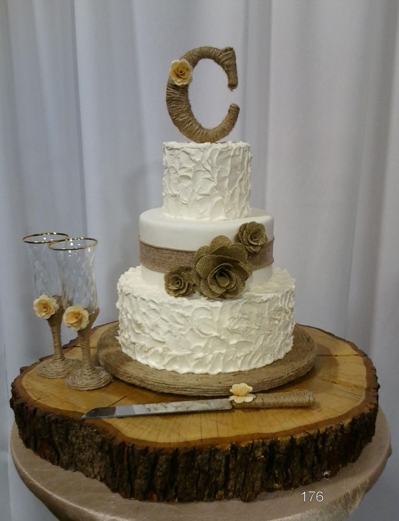 Three-tier white wedding cake with burlap ribbon and gold rose accents on a wood slab, topped with a letter C