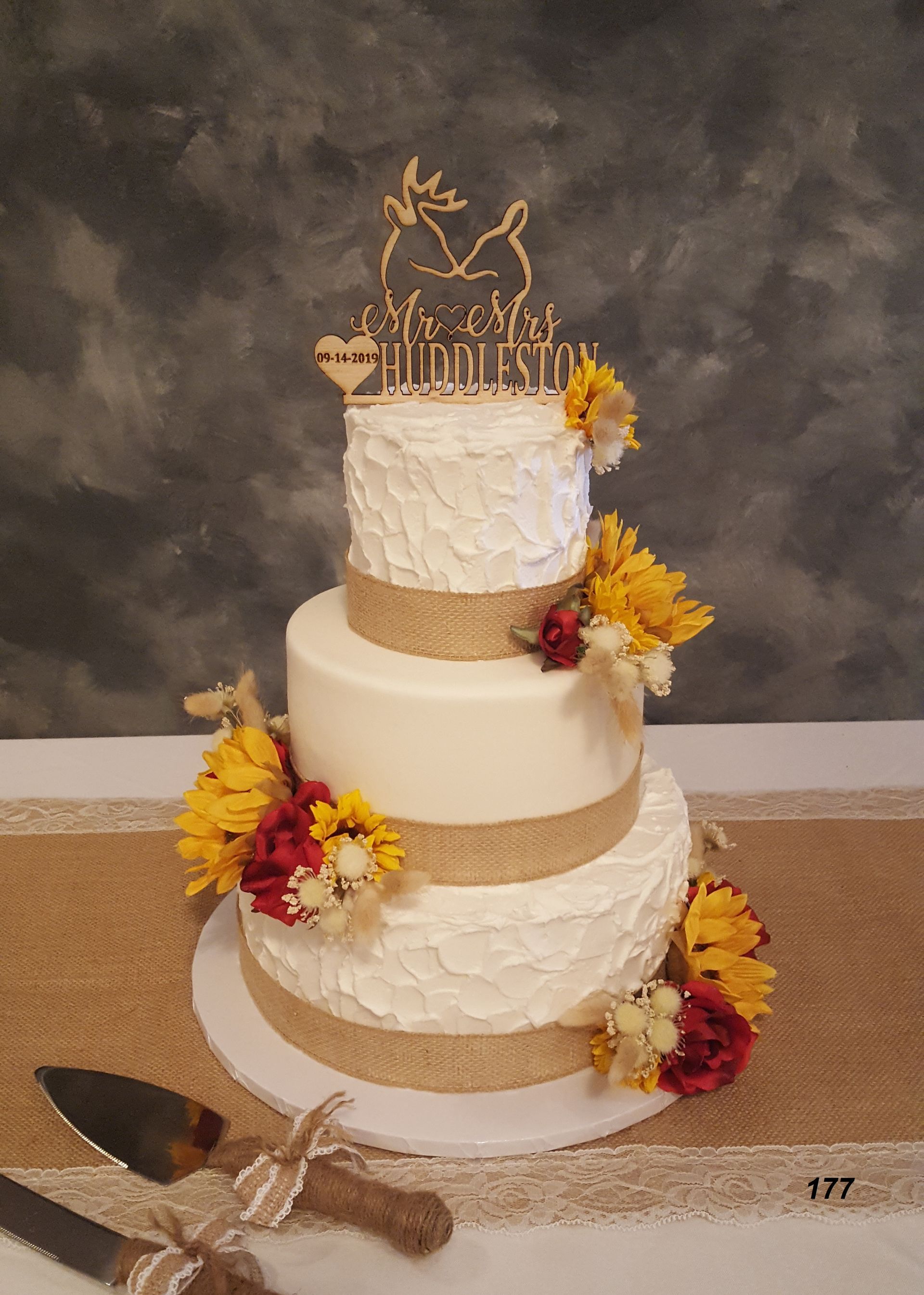 Three-tier white and gold rustic wedding cake with yellow and burgundy flowers and a gold topper