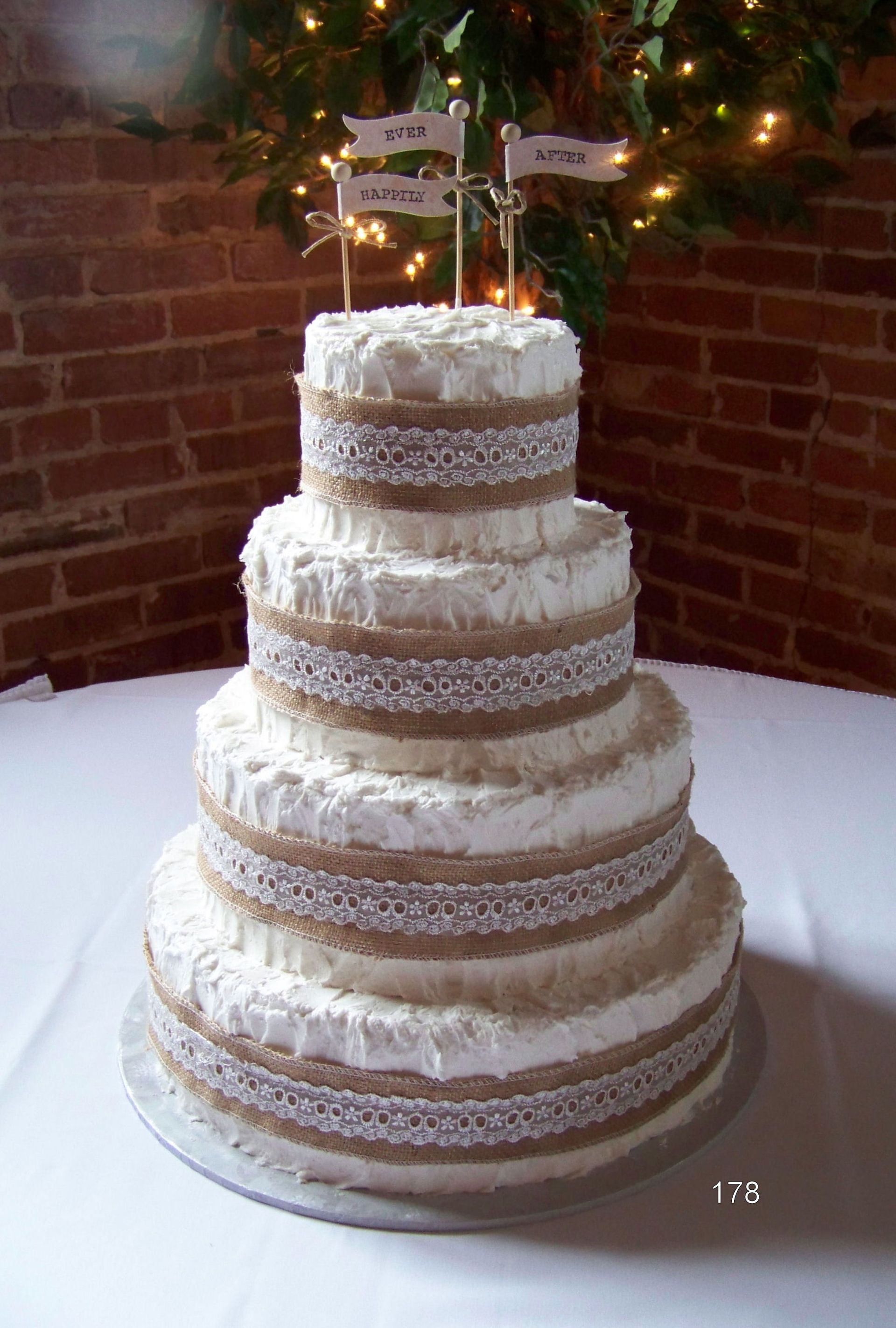 Four-tier white wedding cake with lace trim and small flags on top, on a table with brick wall and lights behind
