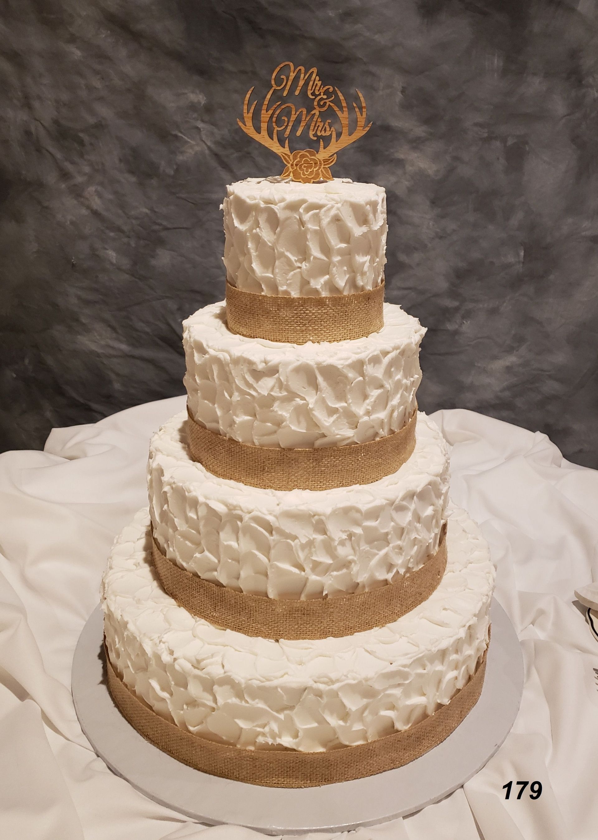 Four-tier white and gold wedding cake with ornate frosting and a gold topper on a white tablecloth.