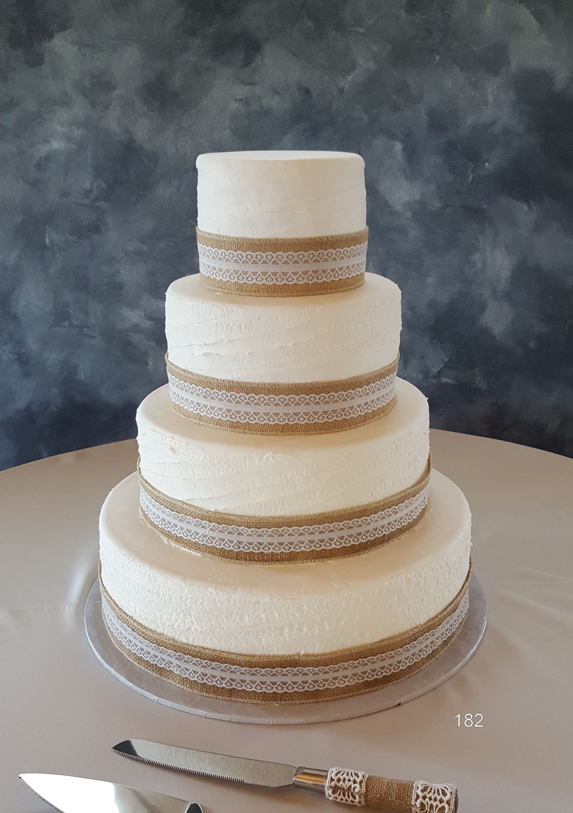 Four-tier ivory wedding cake with lace ribbon accents on a round table, with a knife beside it