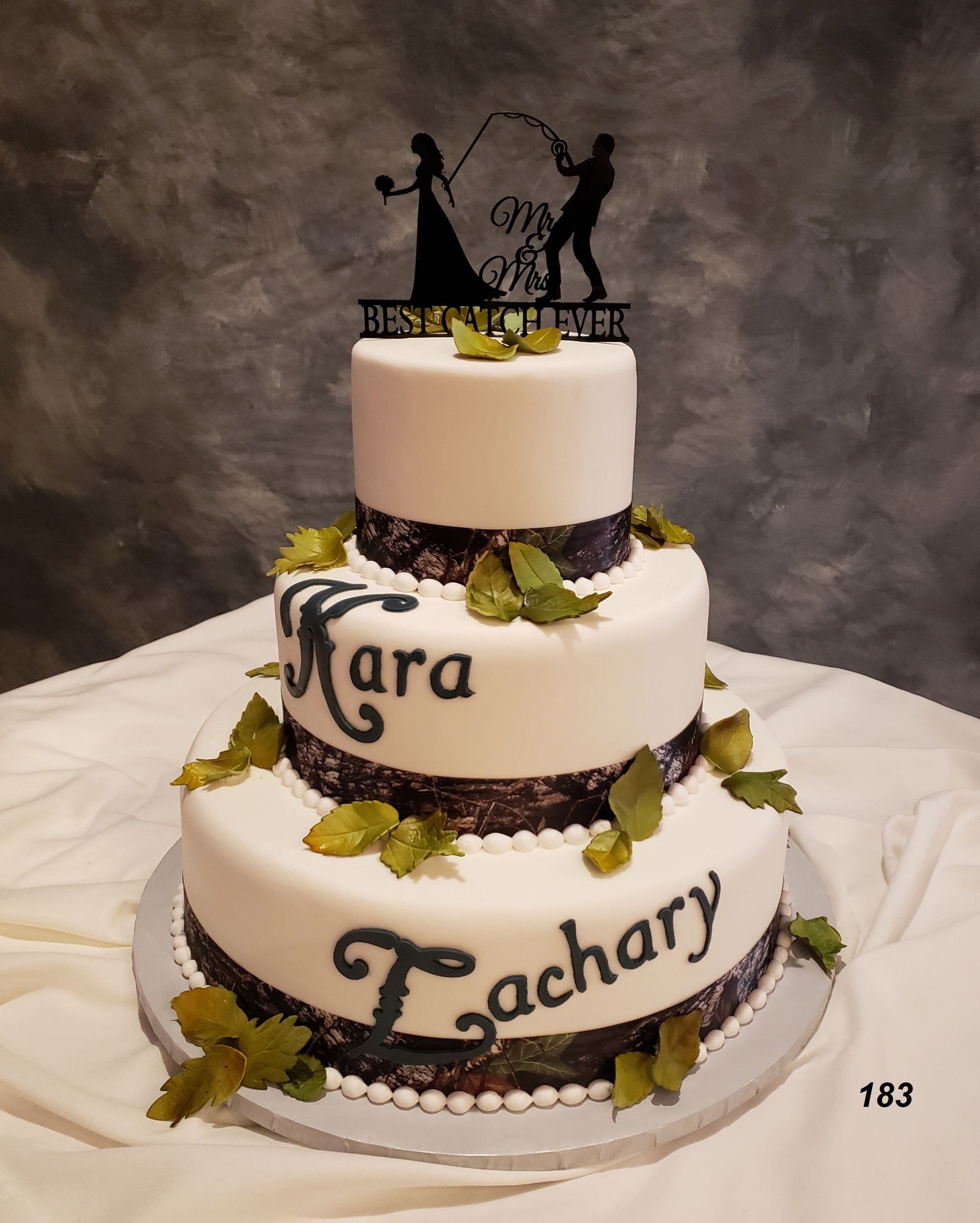 Three-tier white wedding cake with black silhouettes, gold leaves, and “Mara” and “Zachary” lettering.