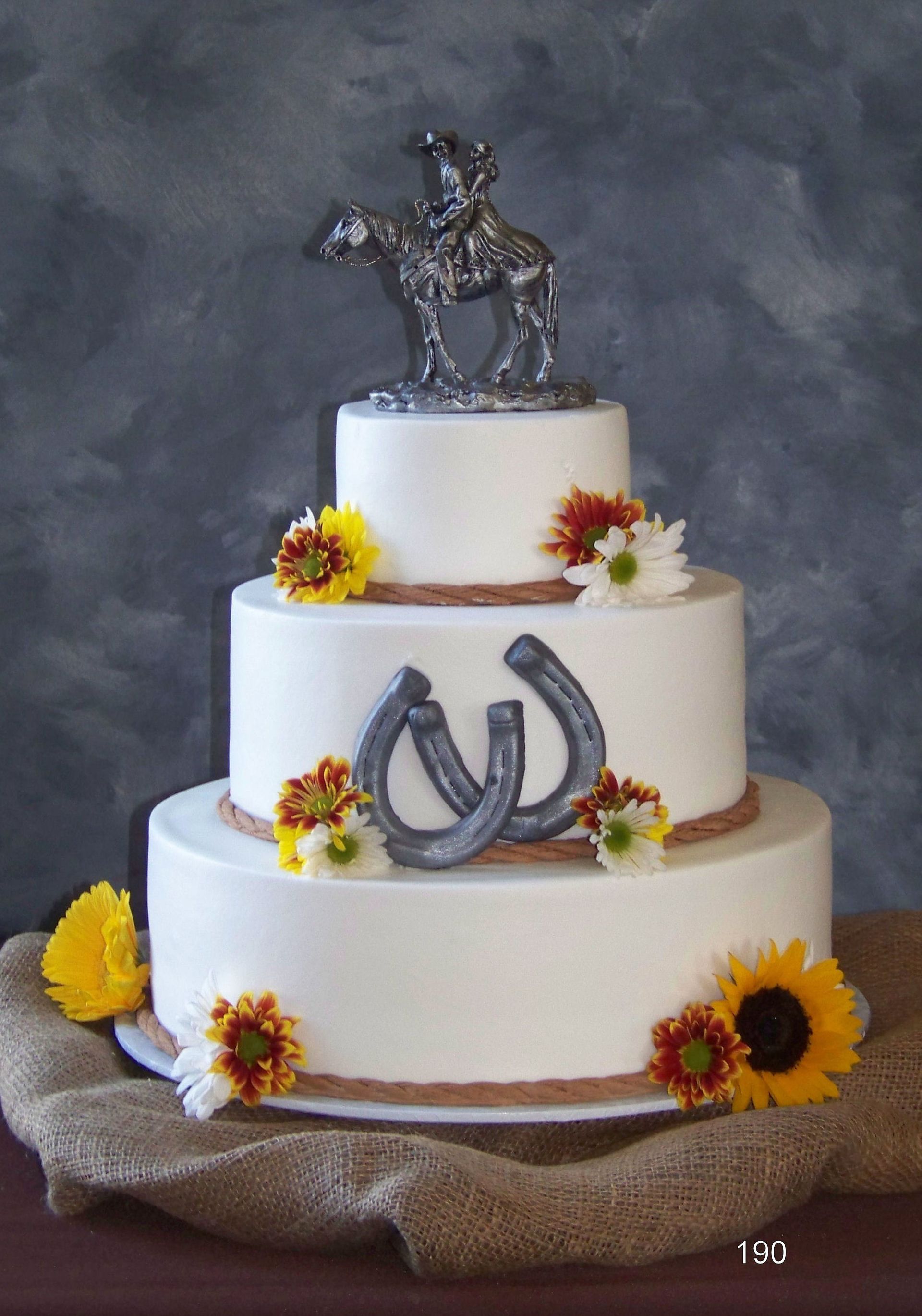 Three-tier white cake with silver horse topper, horseshoe emblem, and yellow-orange flowers on a rustic base
