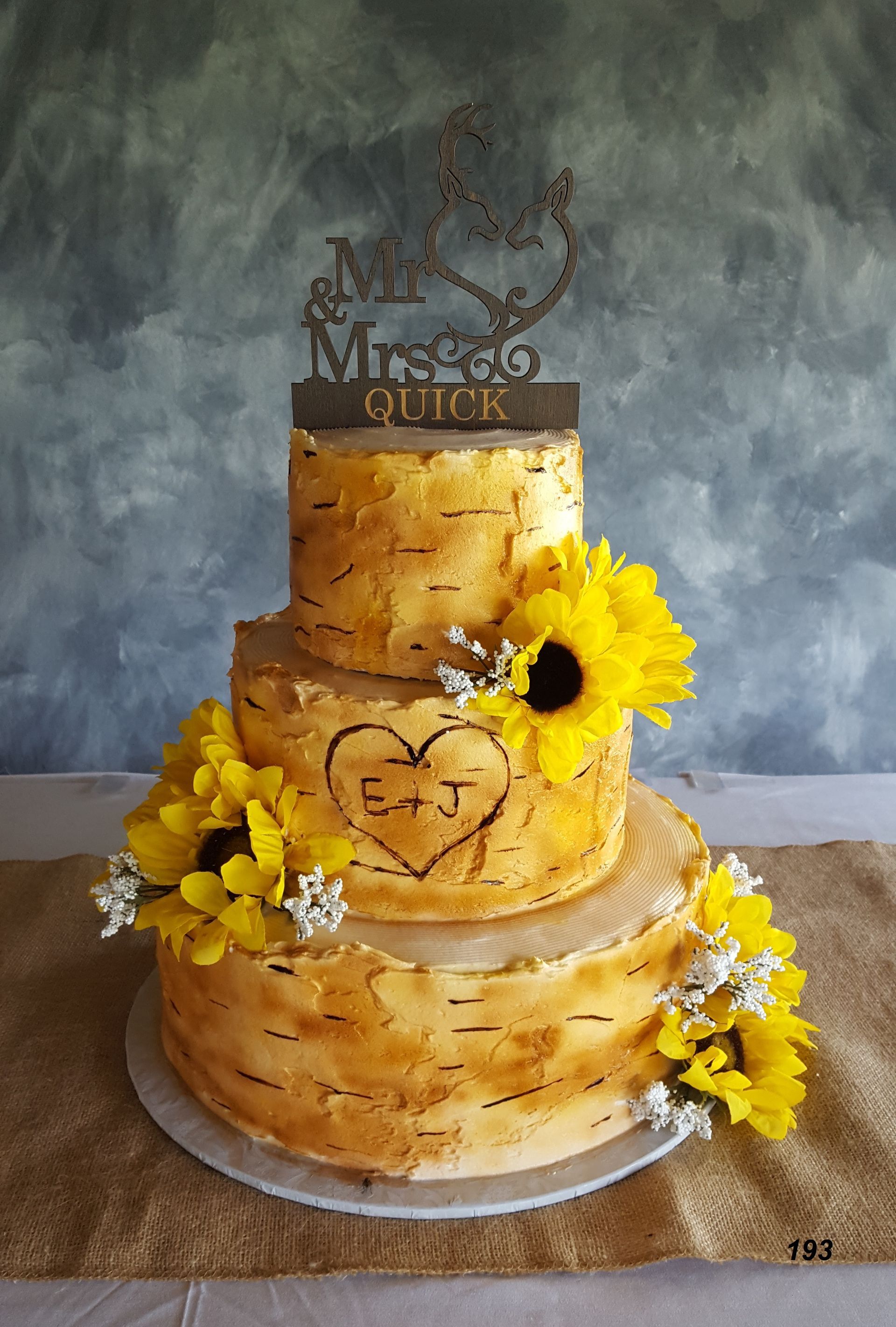 Two-tiered golden cake with yellow flowers and a small topper on a white stand against a blue backdrop