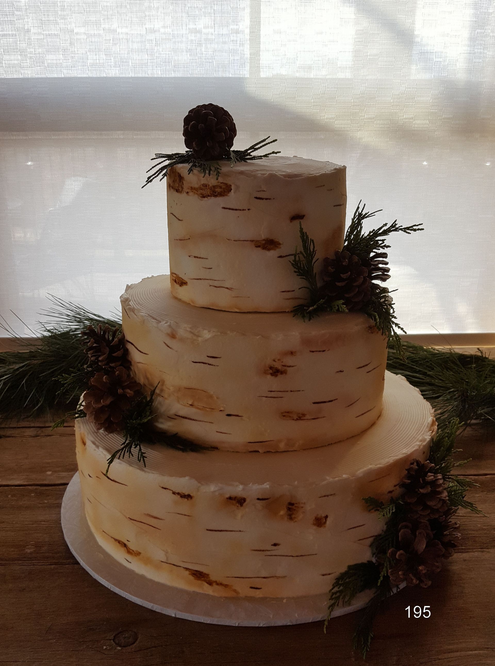 Three-tier white frosted cake with greenery accents on a white plate, backlit by a window