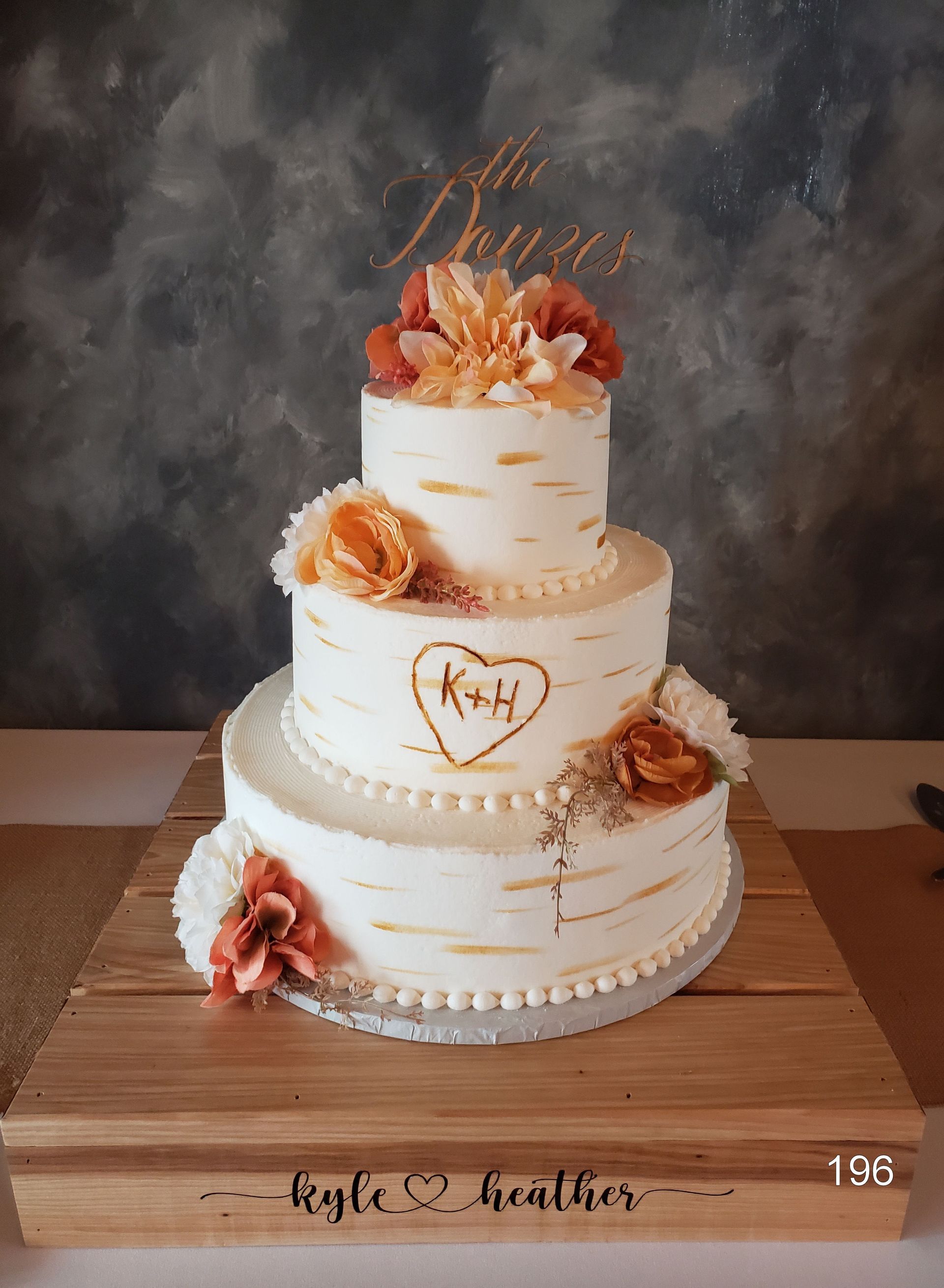Three-tier white wedding cake with peach flowers and gold accents on a wooden table