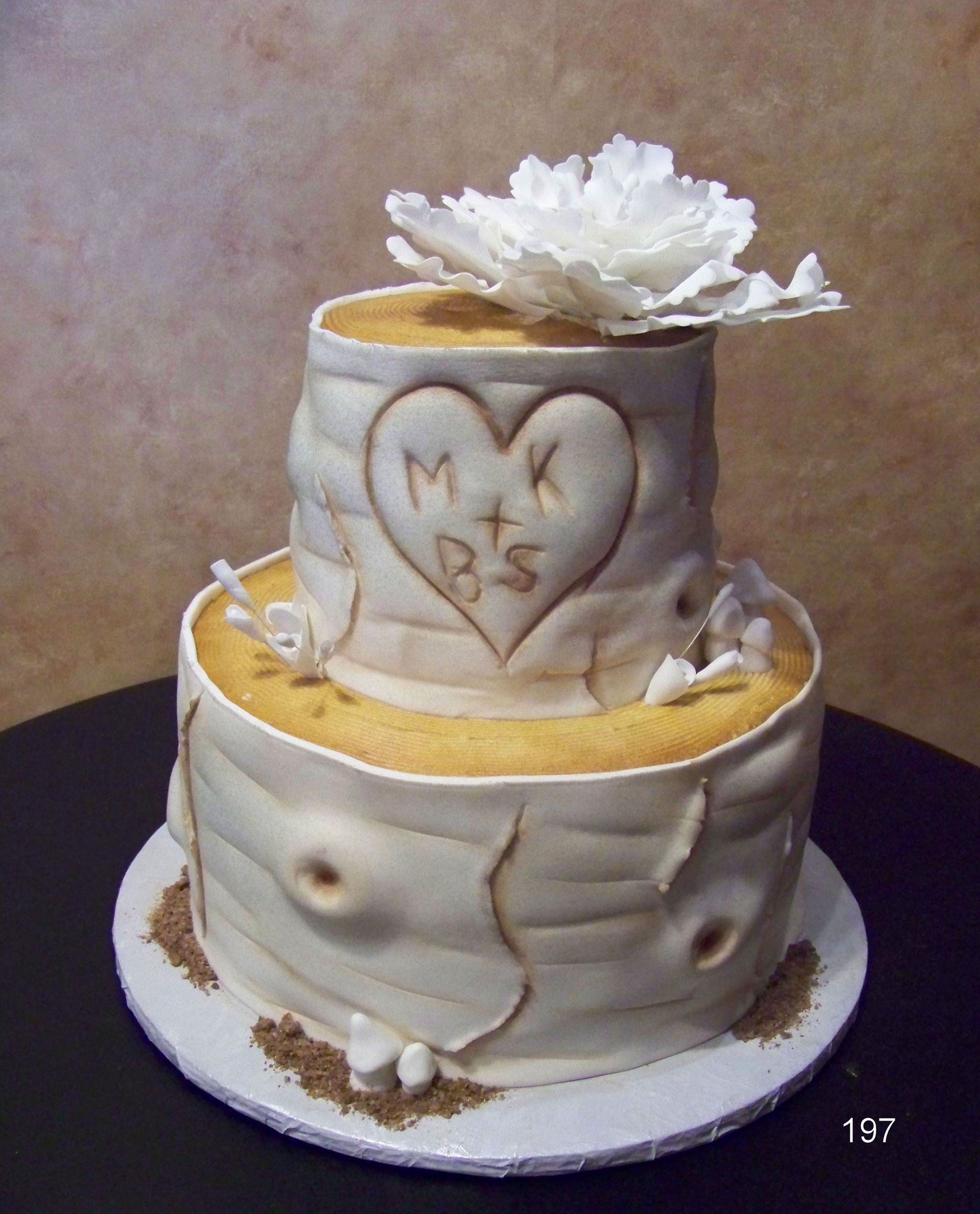 White two-tier decorated cake with a heart and initials on top, topped with a white flower