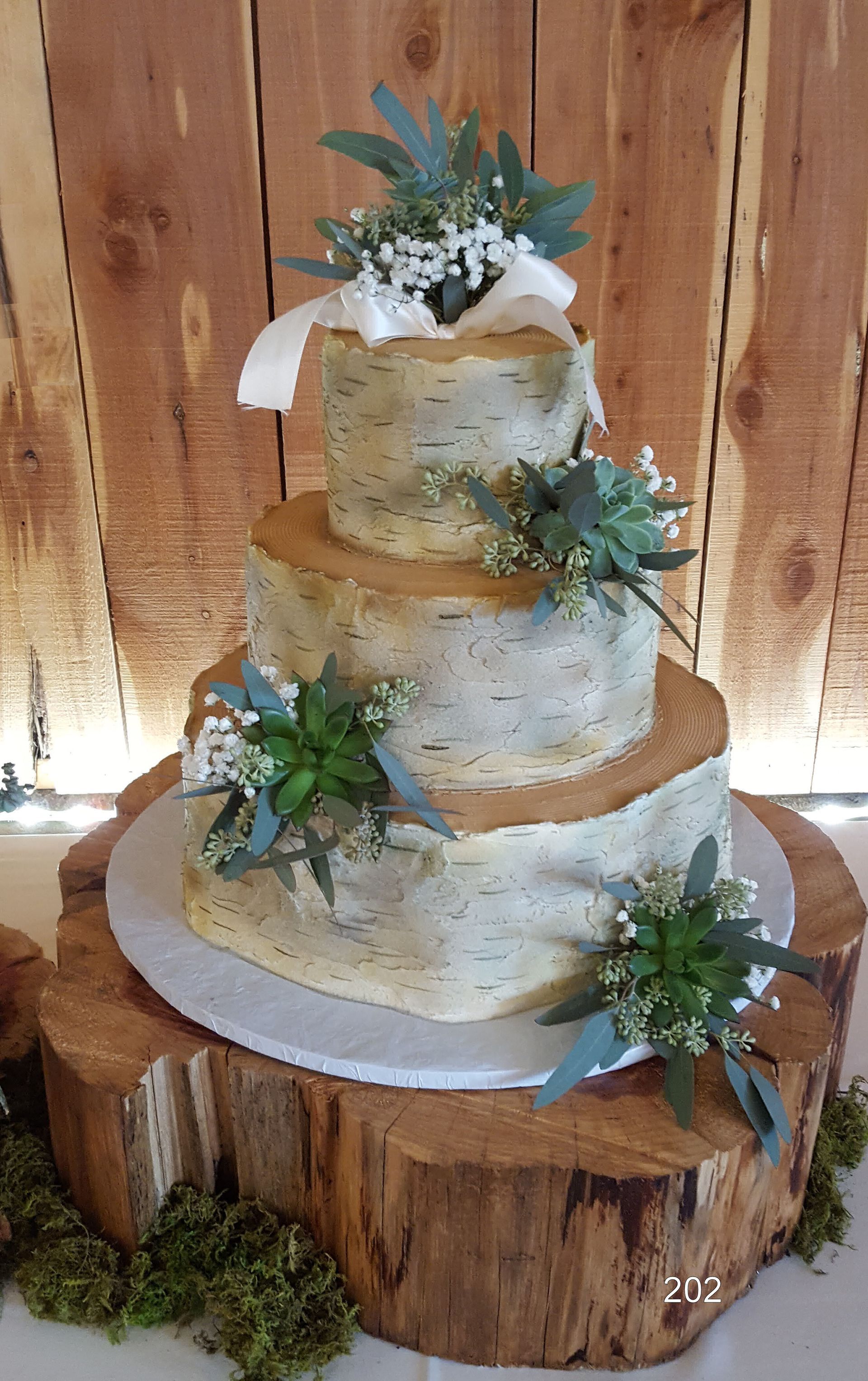 Three-tier white wedding cake with greenery and white flowers on a wooden pedestal, against a wood backdrop