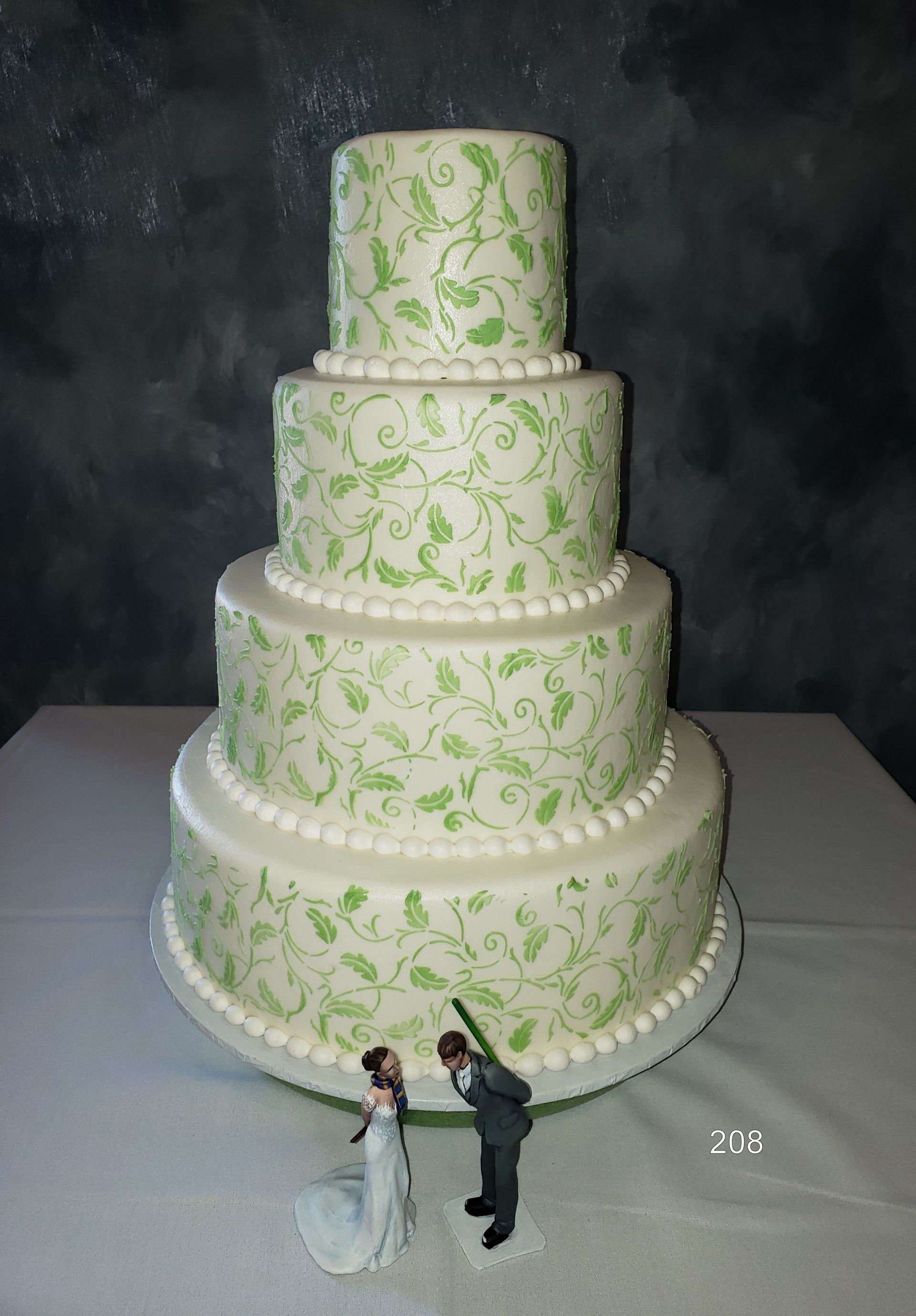 Four-tier white wedding cake with green vines and bride and groom figurines at the base