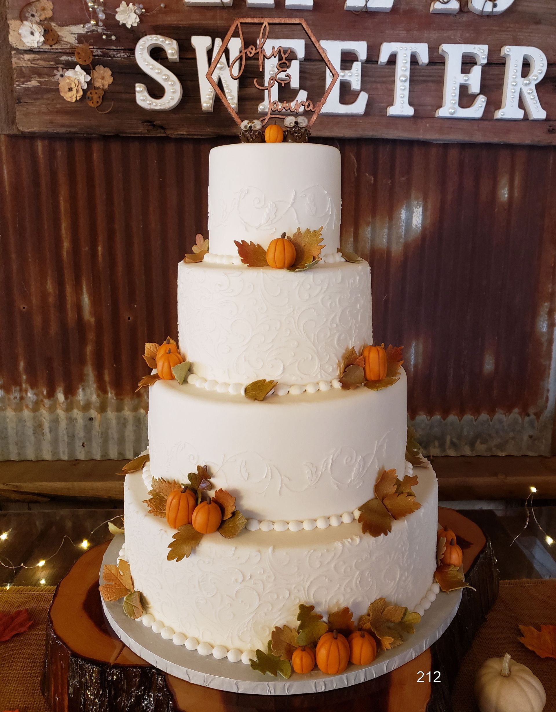 Four-tier white cake with orange flowers and pumpkins under a “SWEETER” sign