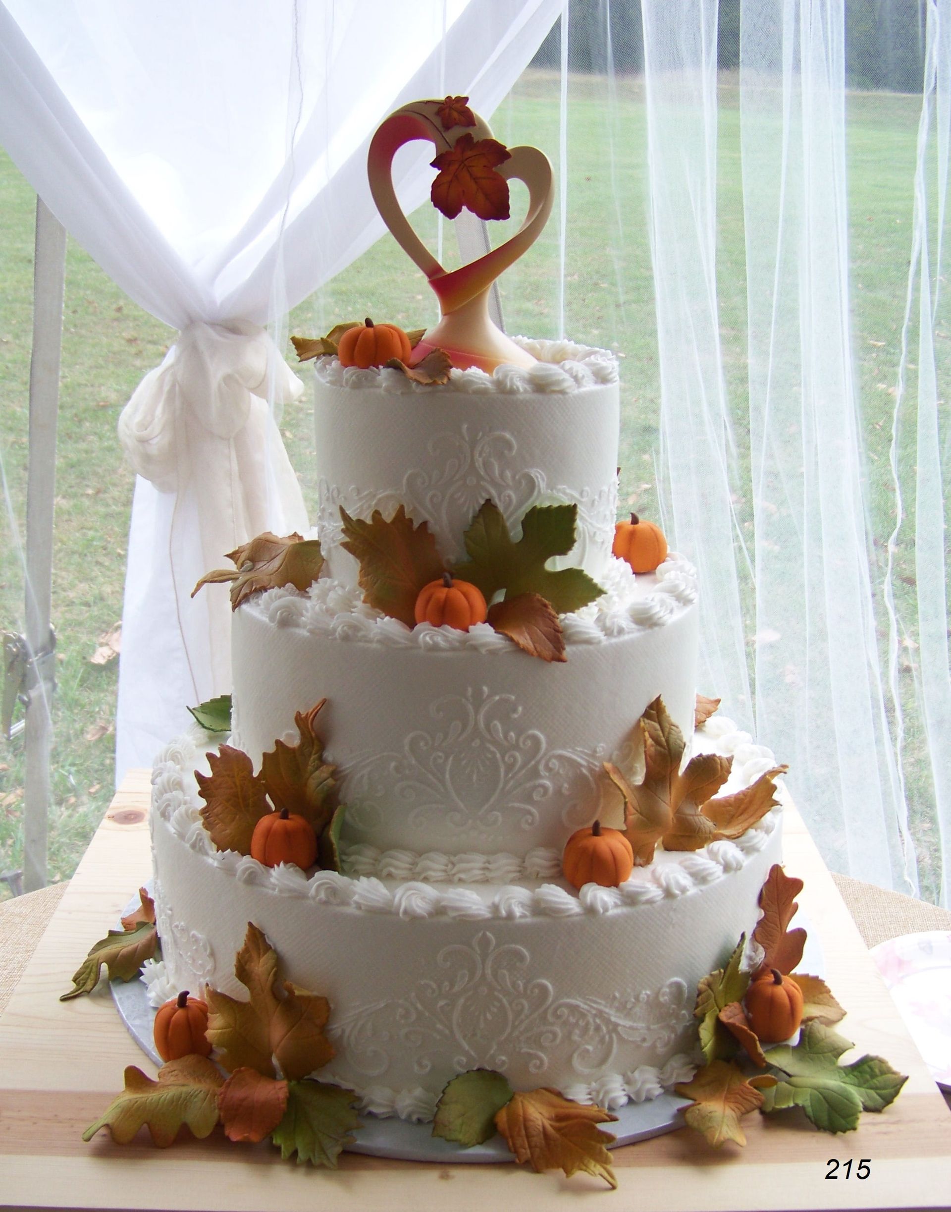 Three-tier white wedding cake with autumn leaves and a heart topper by a window