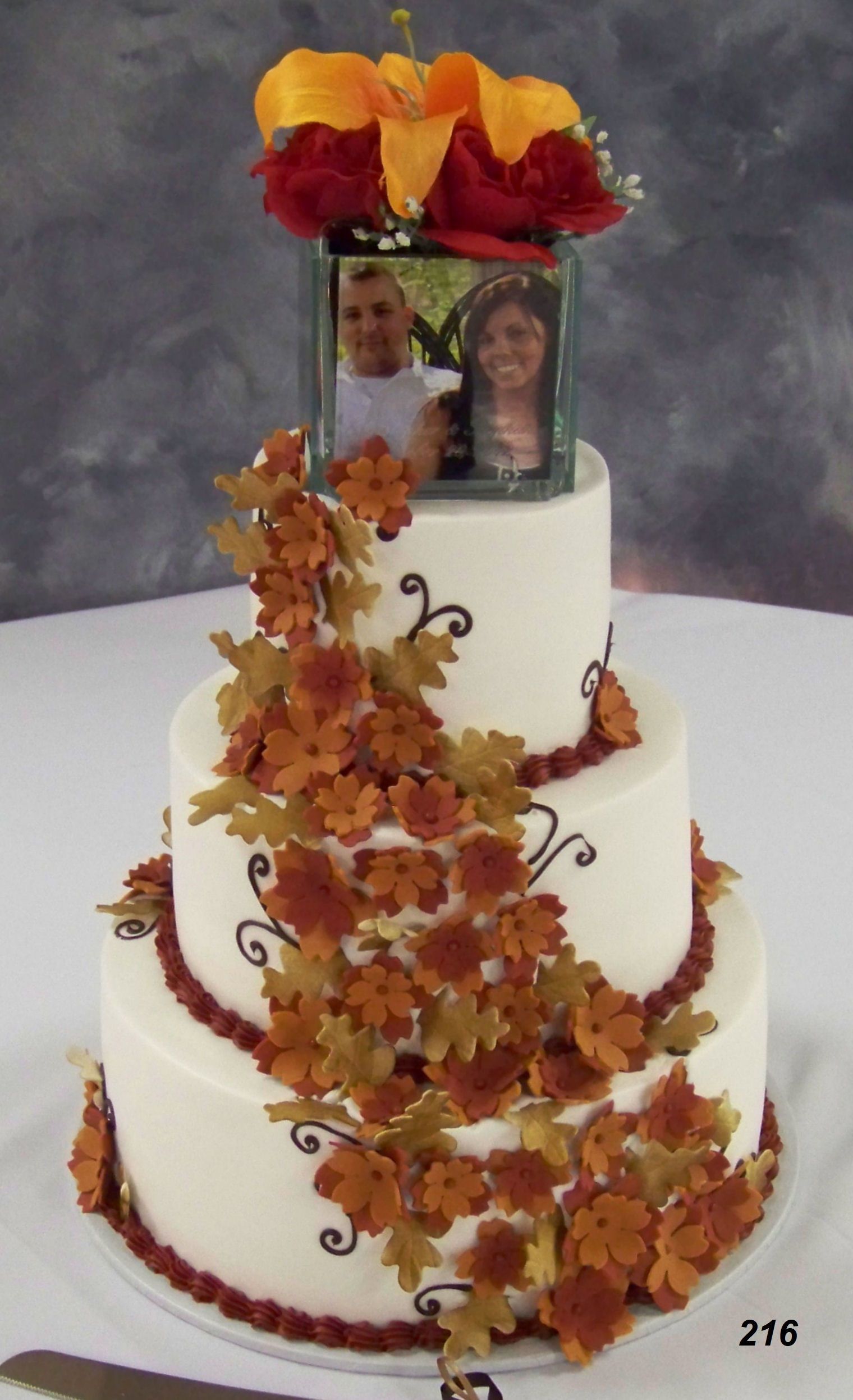 Three-tier white cake decorated with autumn leaves and two framed photos on top, topped with orange and red flowers