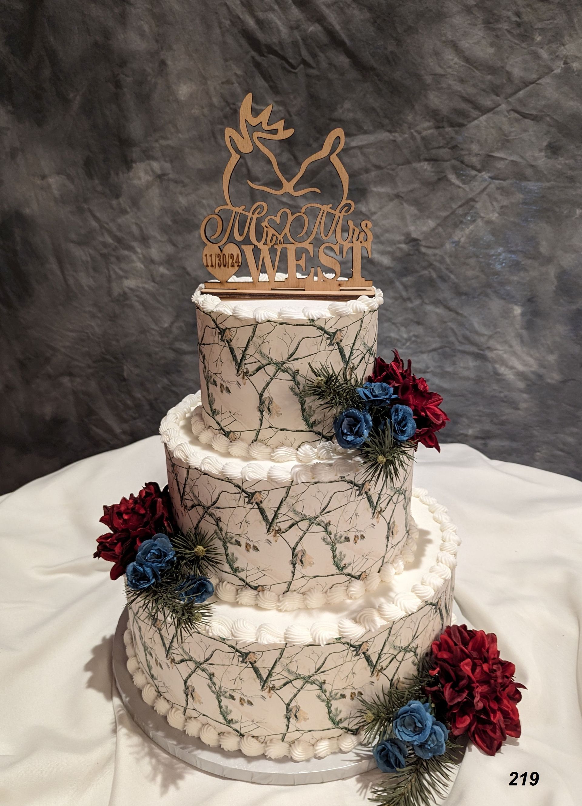 Three-tier white wedding cake with gold topper and red and blue flowers on a gray backdrop