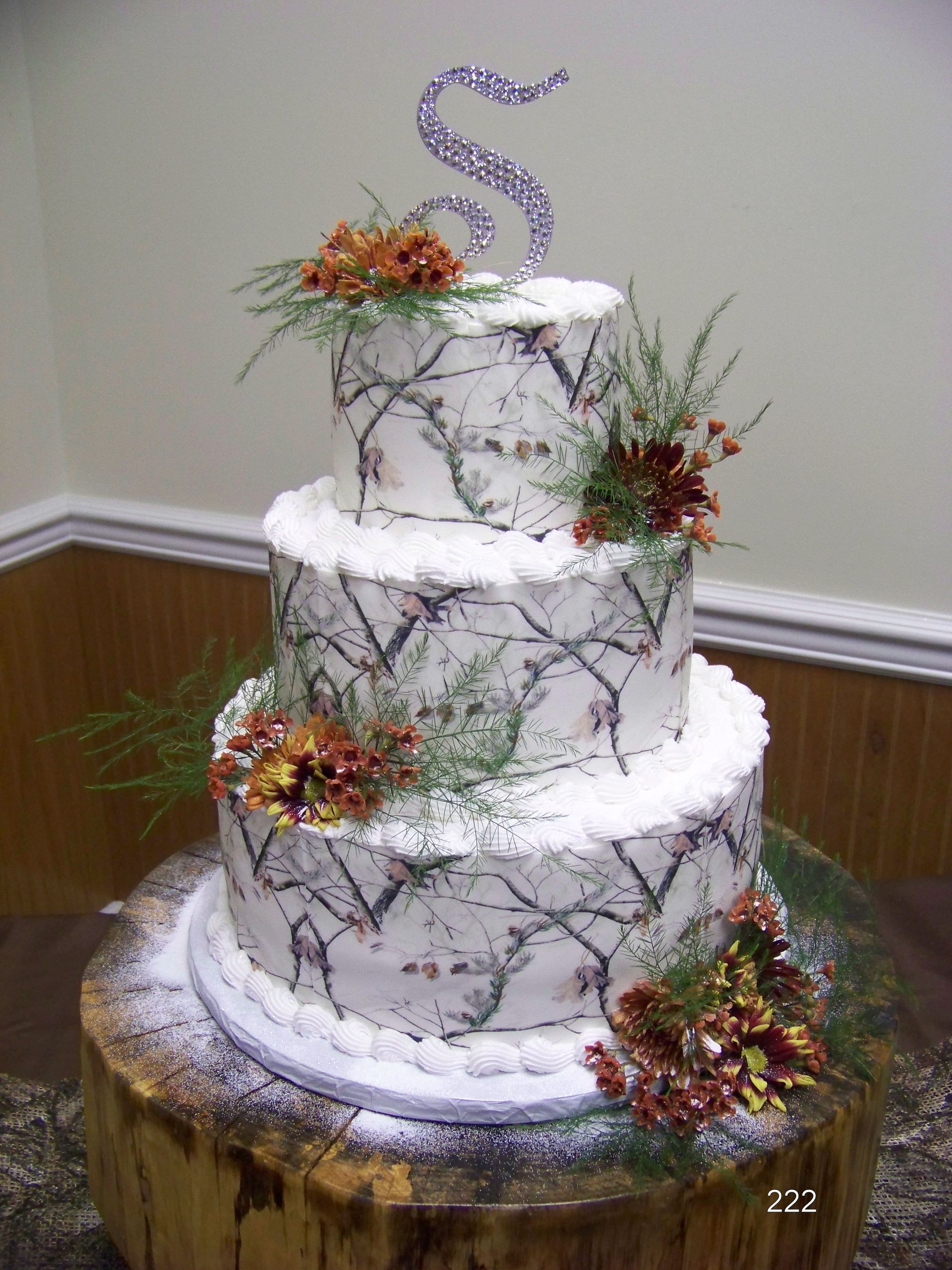 Three-tier white wedding cake with floral decorations and a silver monogram topper on a wooden table