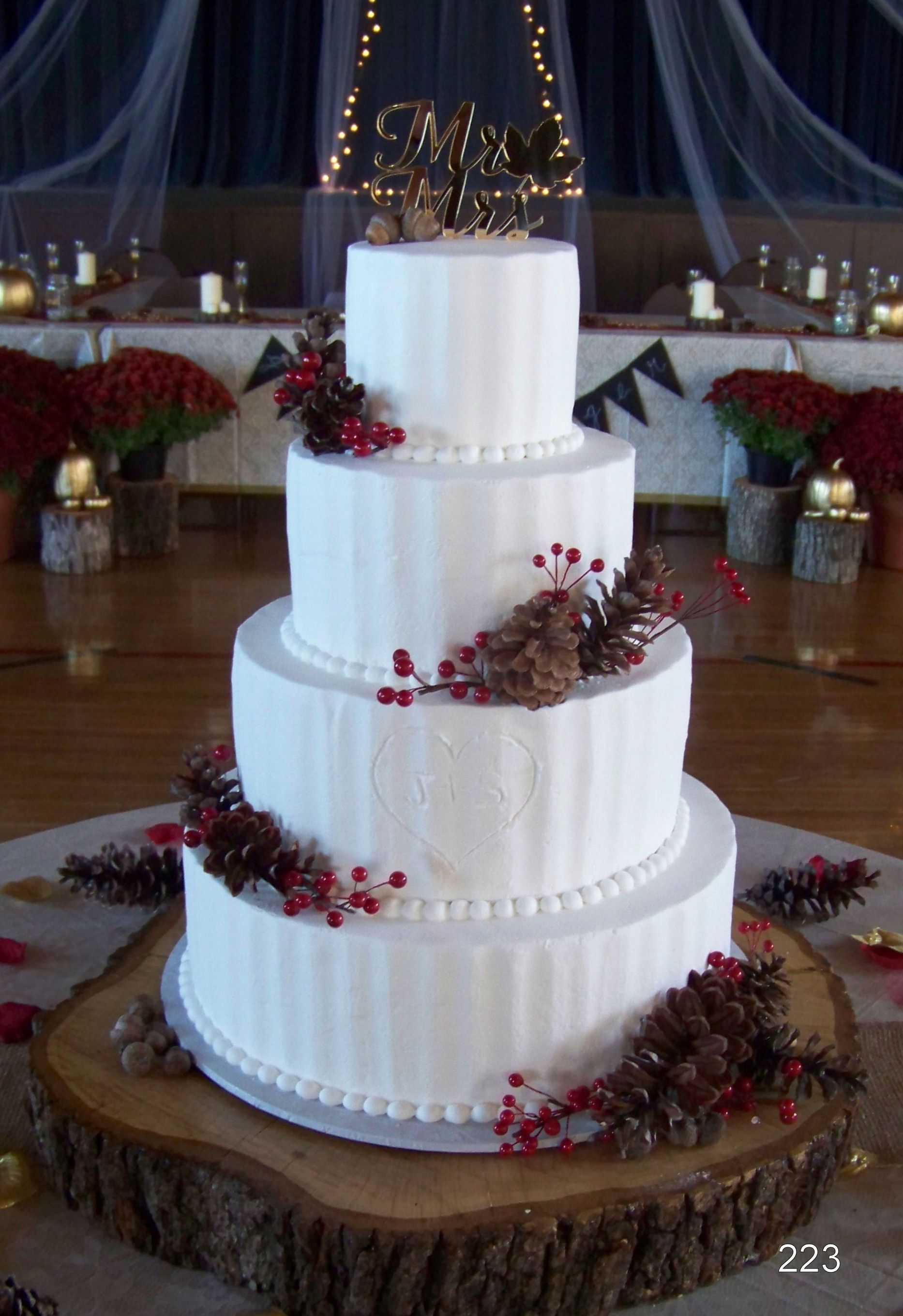 Four-tier white wedding cake with red berry and pinecone decorations on a rustic wood slab table