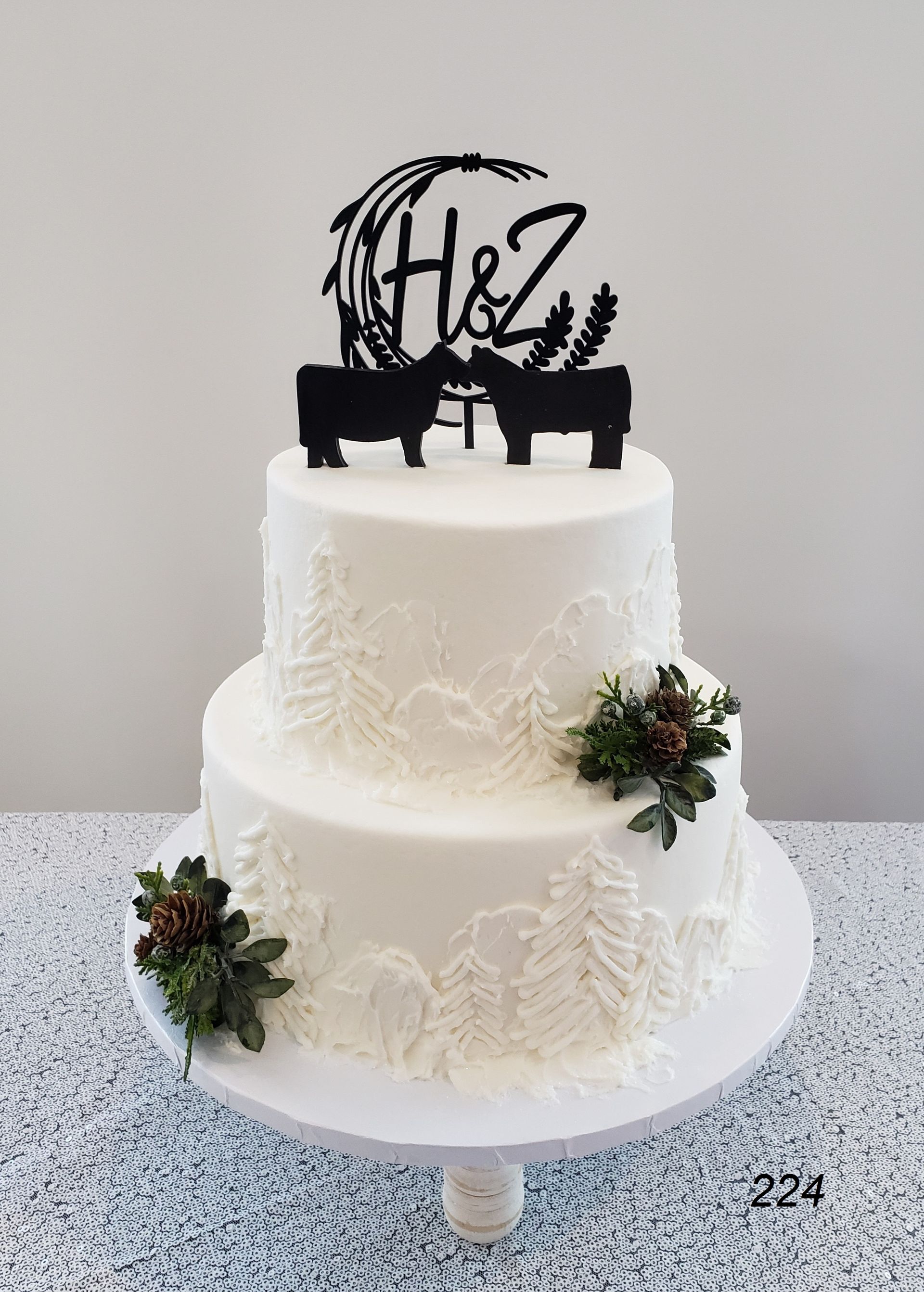 White two-tier cake with black cow toppers and floral decorations on a stand