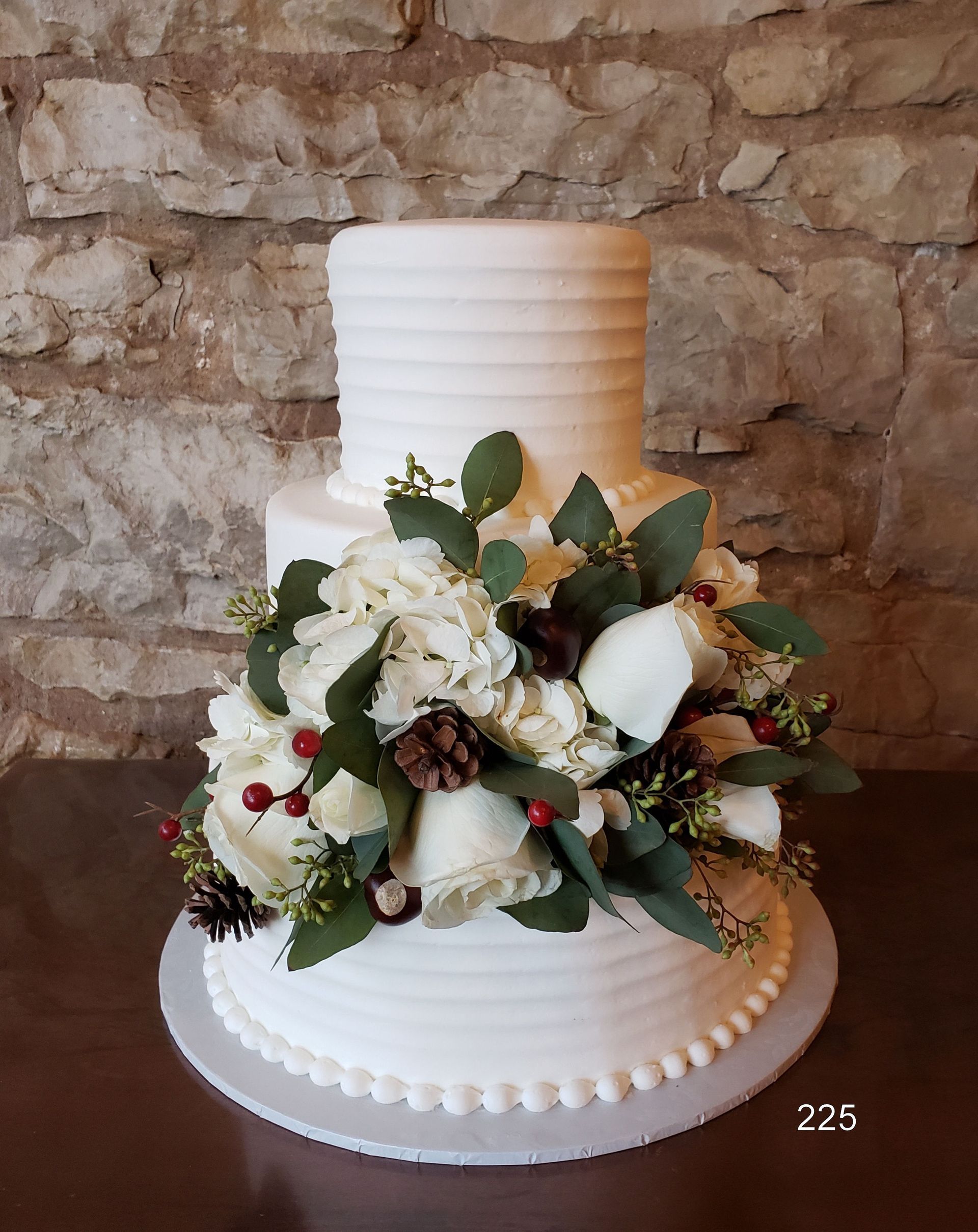White tiered cake with white flowers, green leaves, and red berries on a stand against a stone wall