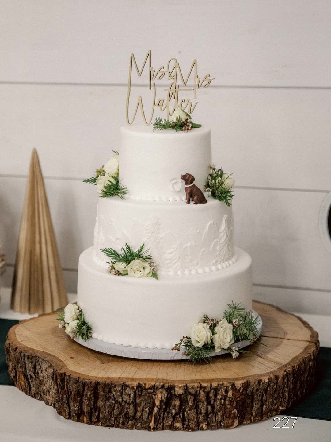 Three-tier white wedding cake with gold topper, white flowers, and a small couple figurine on a wood slab.