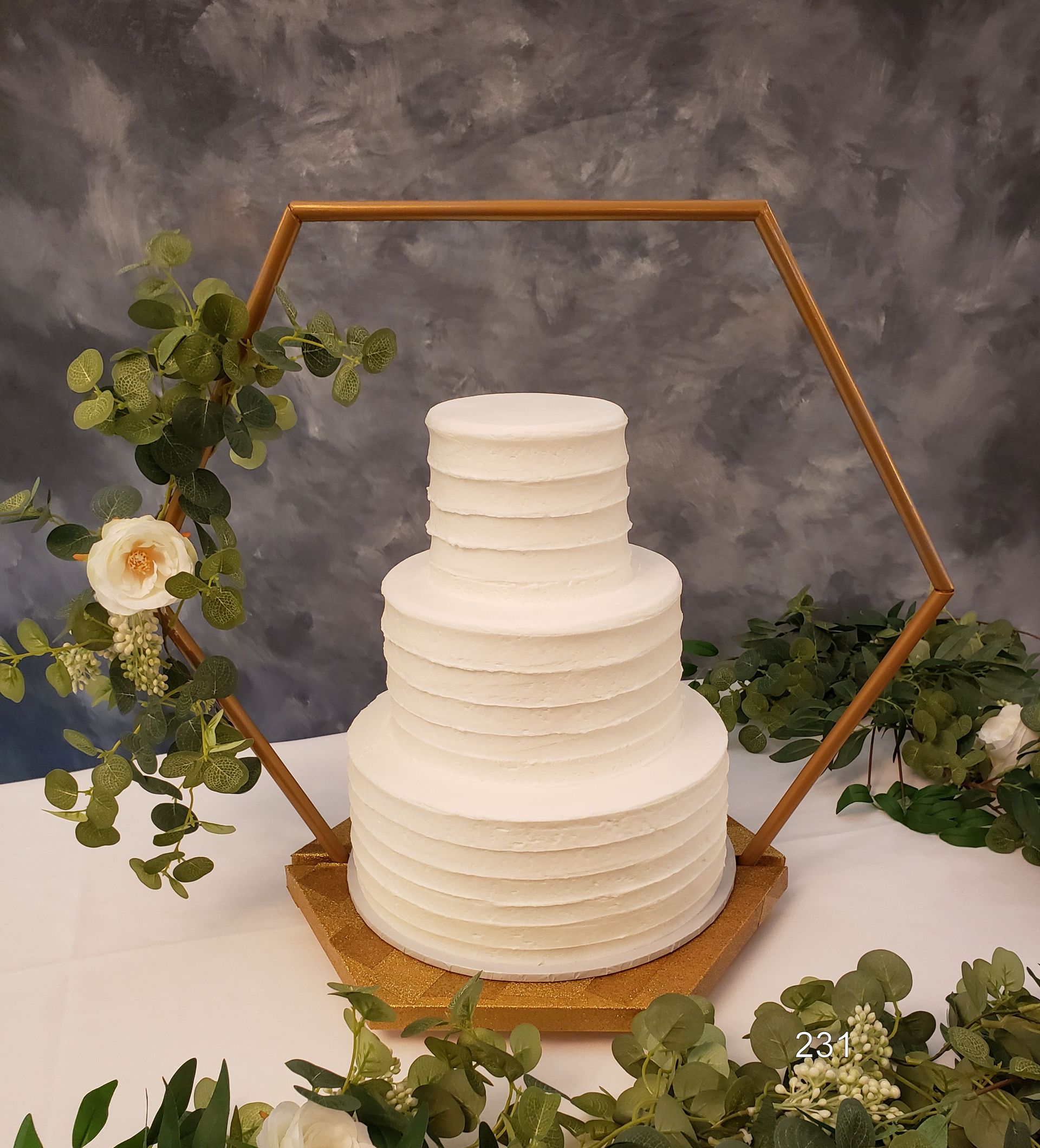 White tiered cake on a gold hexagonal stand with greenery and flowers against a gray backdrop