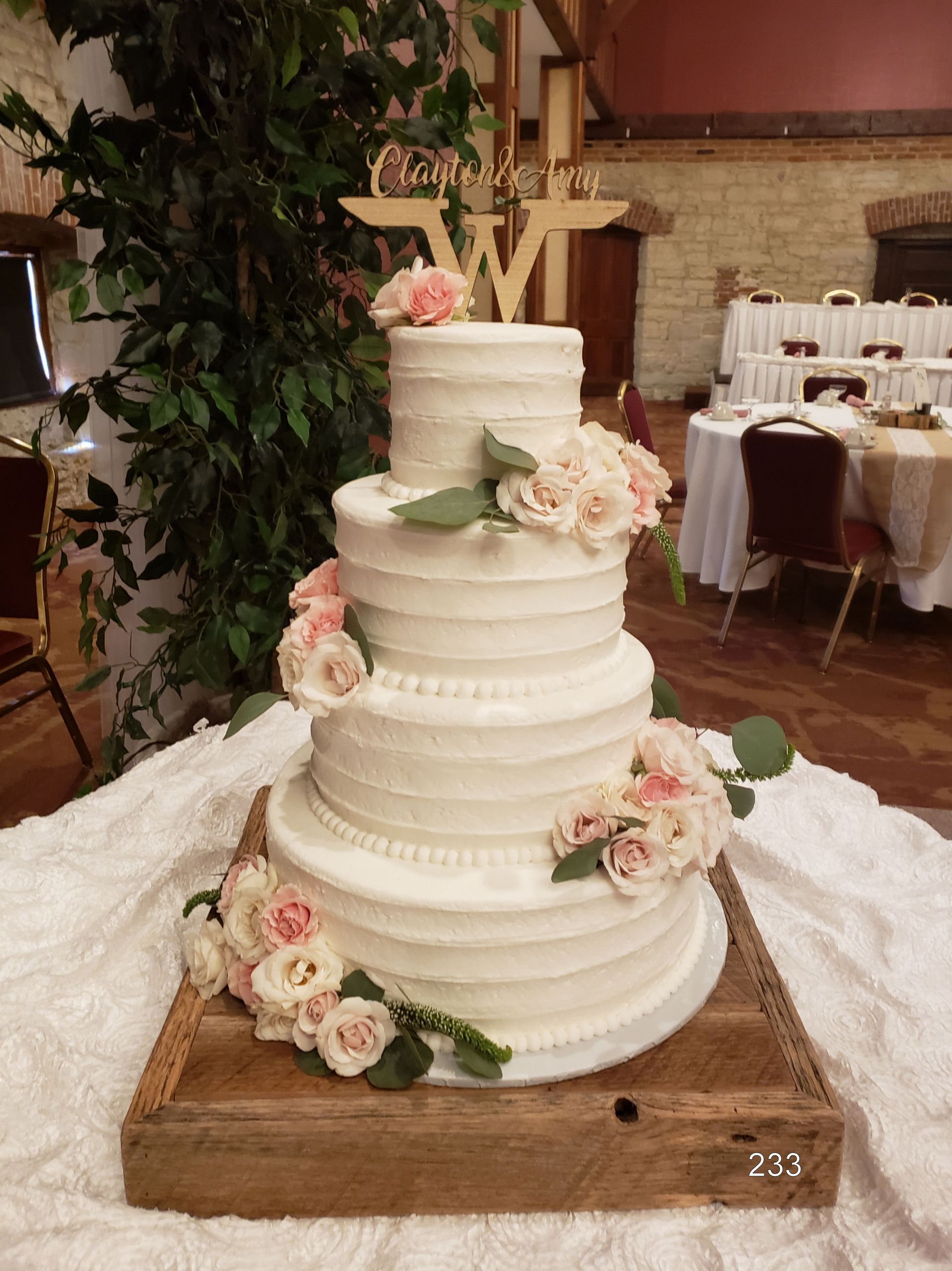 White tiered wedding cake with pink flowers and greenery on a wooden stand at a reception hall