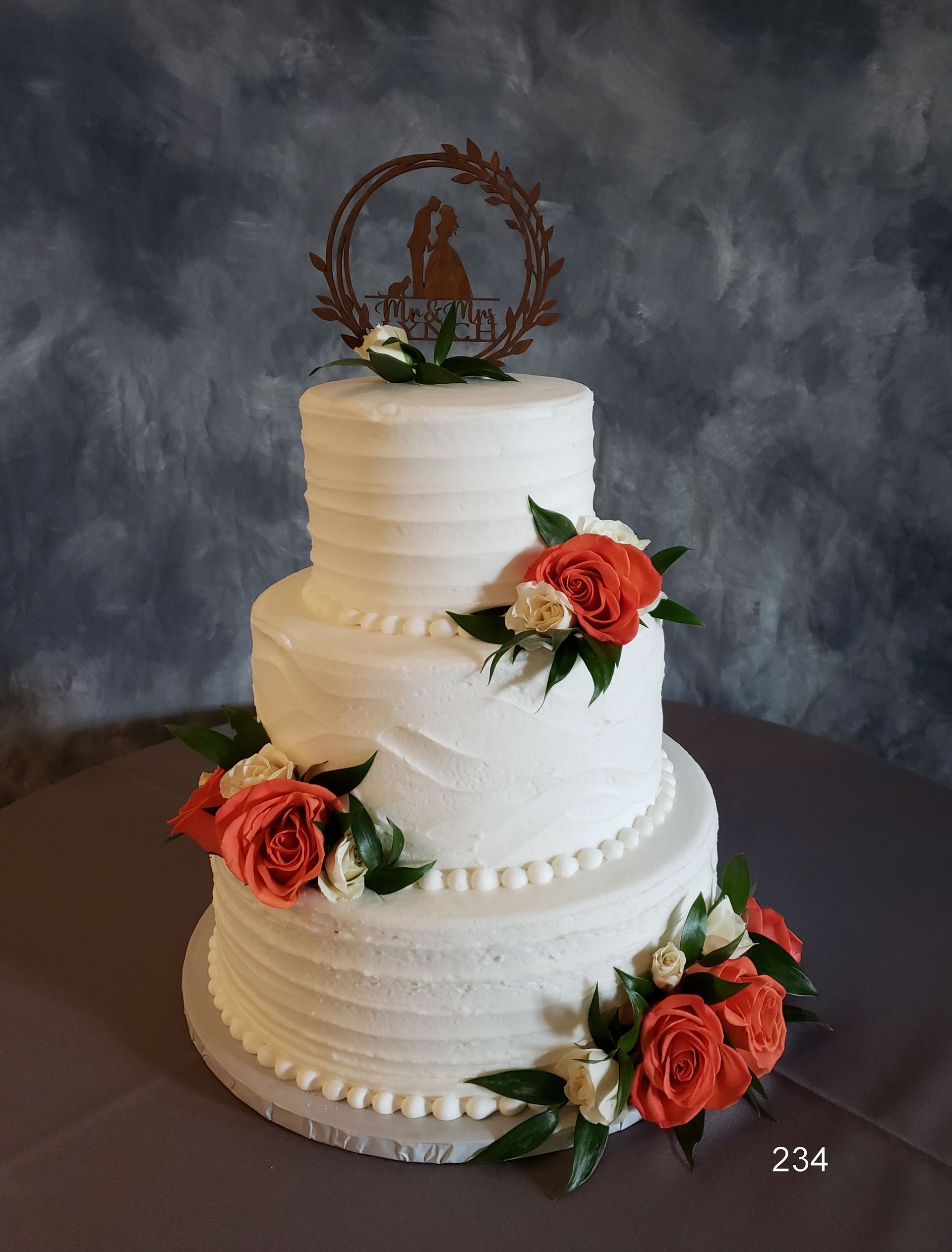Three-tier white wedding cake with coral roses and a rustic topper on a dark backdrop