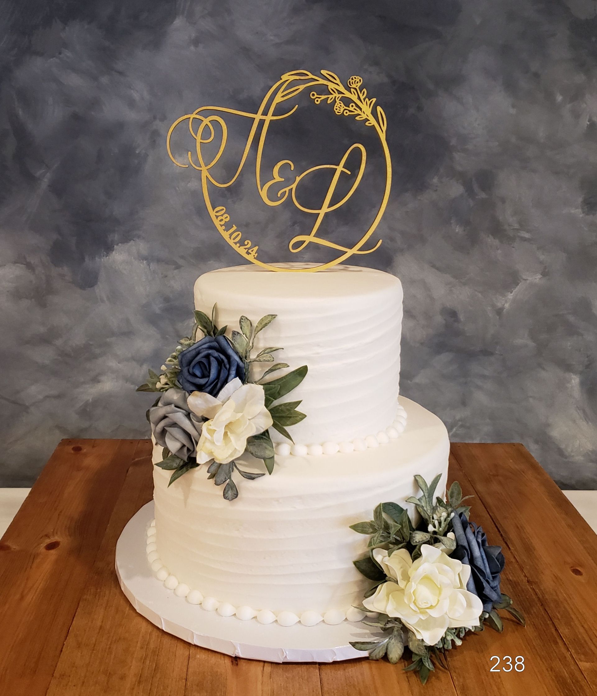 Two-tier white wedding cake with navy and cream flowers and a gold monogram topper on a wooden table
