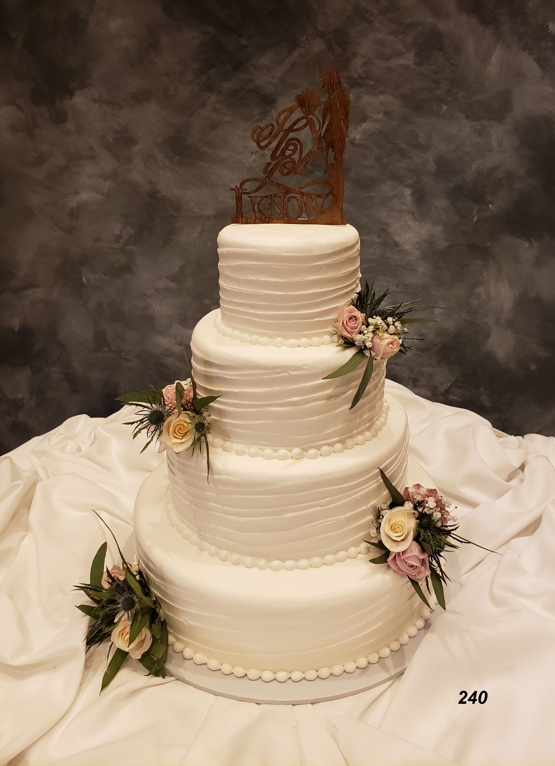 Four-tier white wedding cake with pastel floral accents and a decorative topper on a draped table.