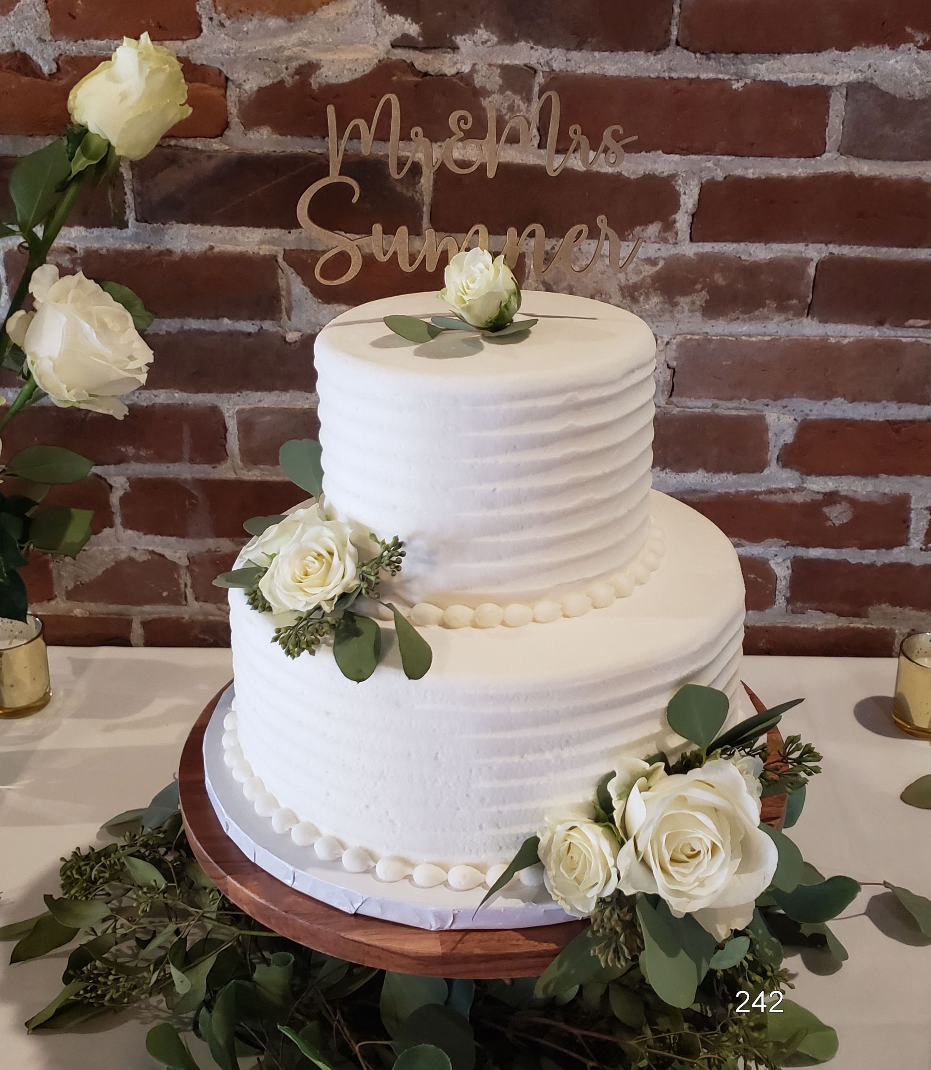 Two-tier white wedding cake with white flowers and gold topper against a brick wall