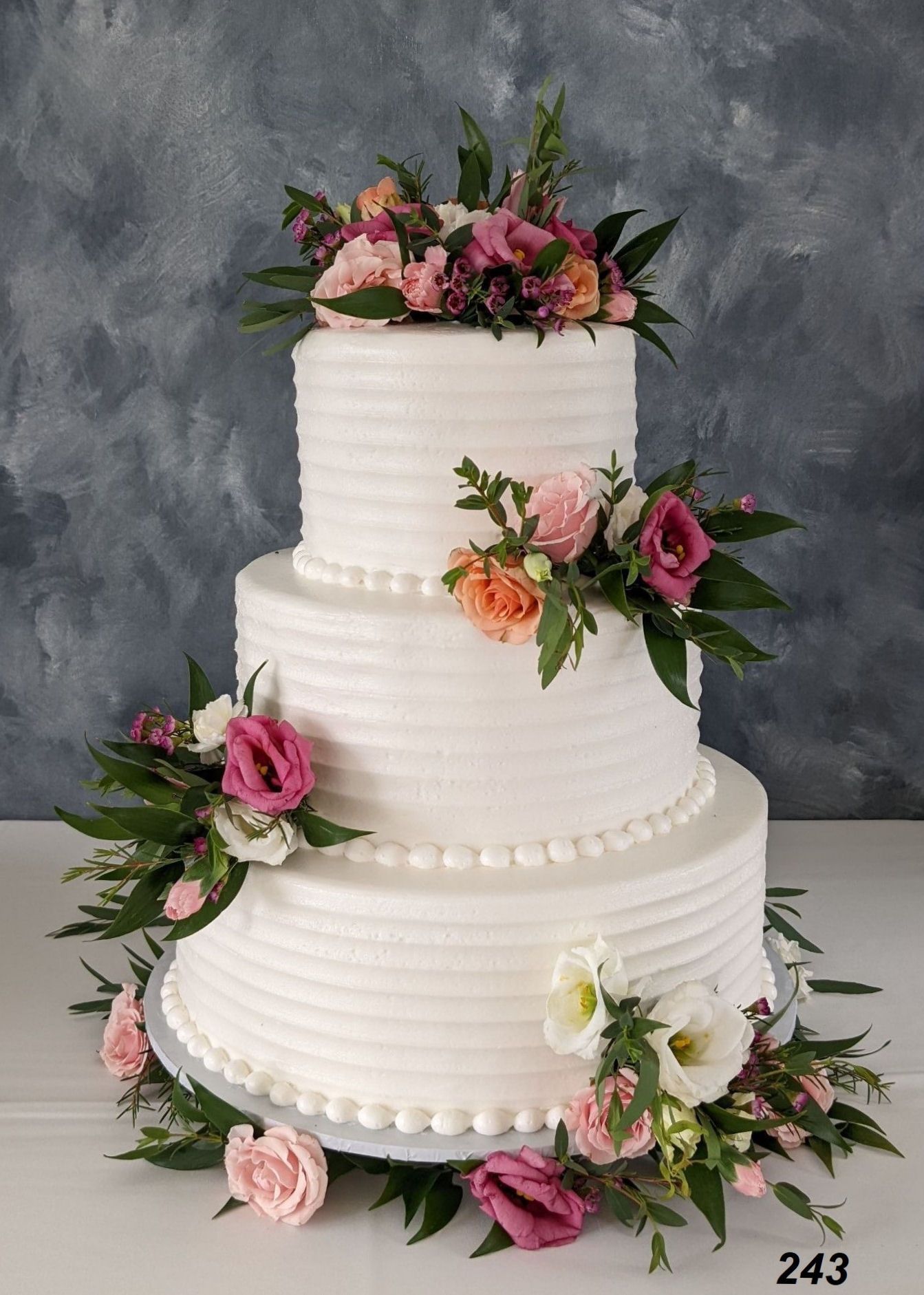 Three-tier white wedding cake with pink floral decorations on a gray backdrop