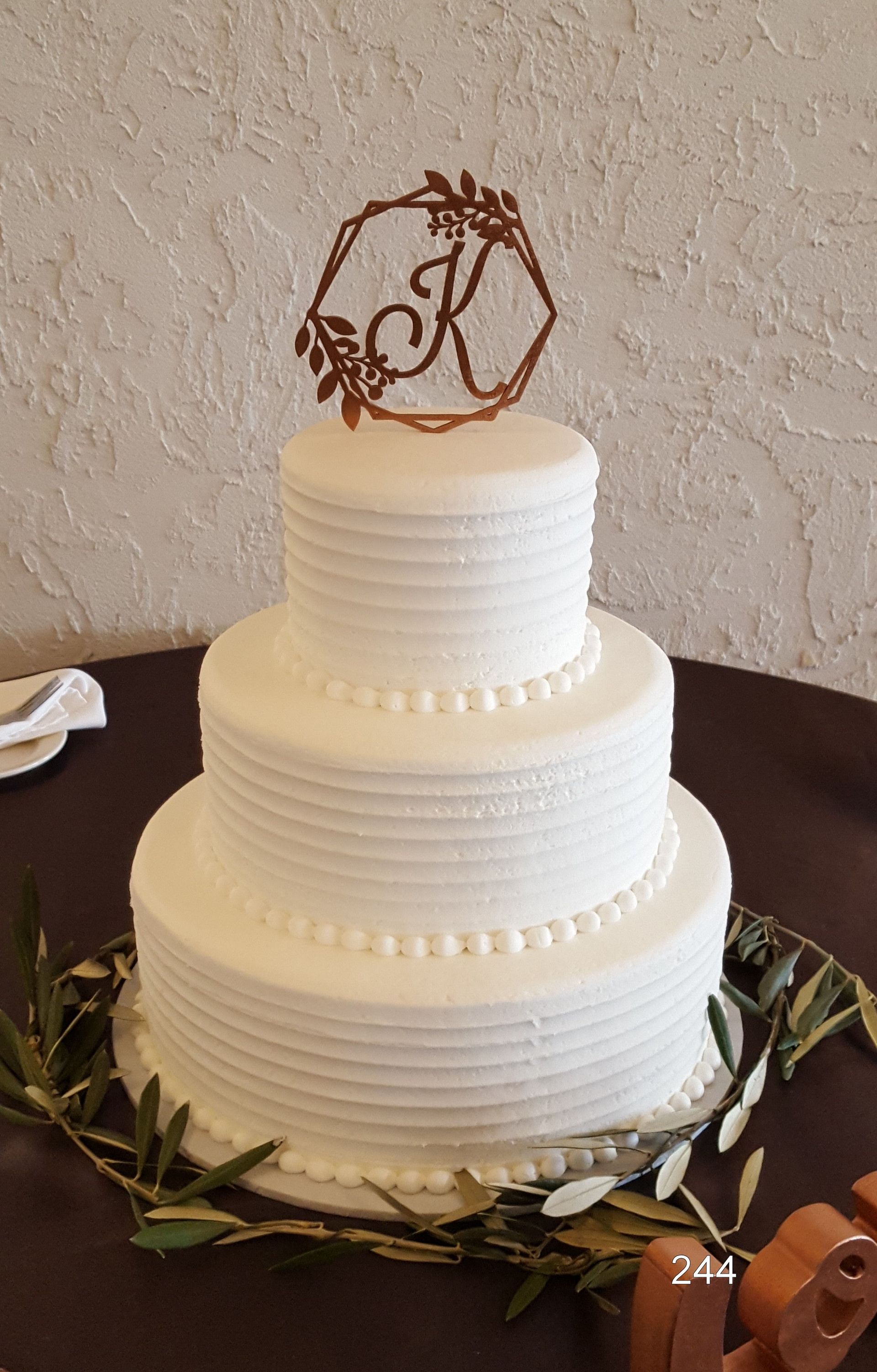 Three-tier white wedding cake with gold monogram topper and greenery on the base