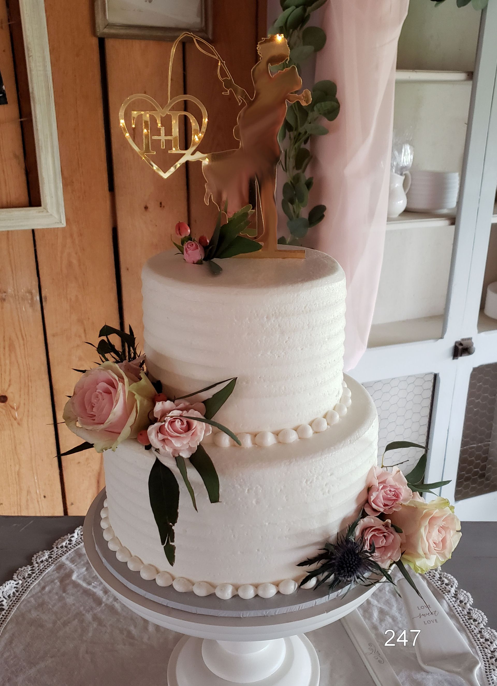 Two-tier white wedding cake with pink roses and gold heart topper on a stand