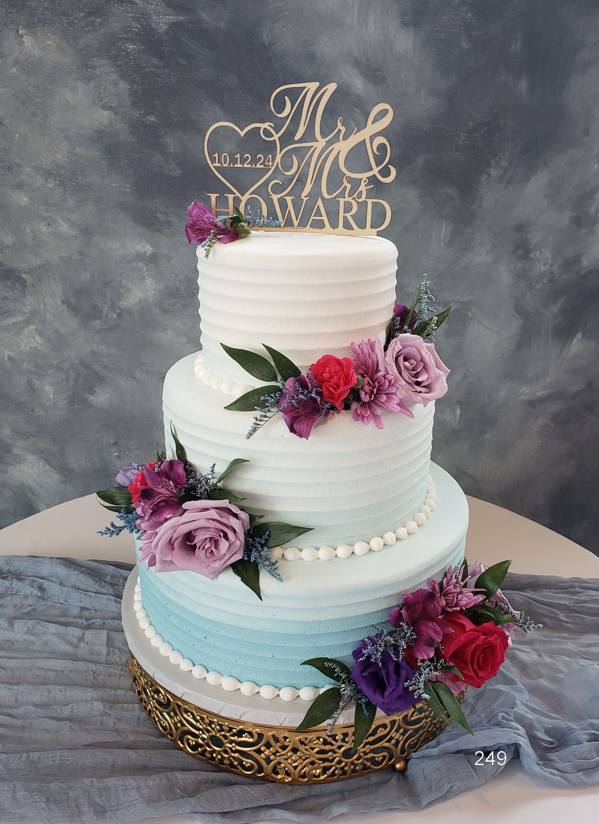 Three-tier white cake with pink and purple flowers, gold topper, and gold lattice base on a gray backdrop