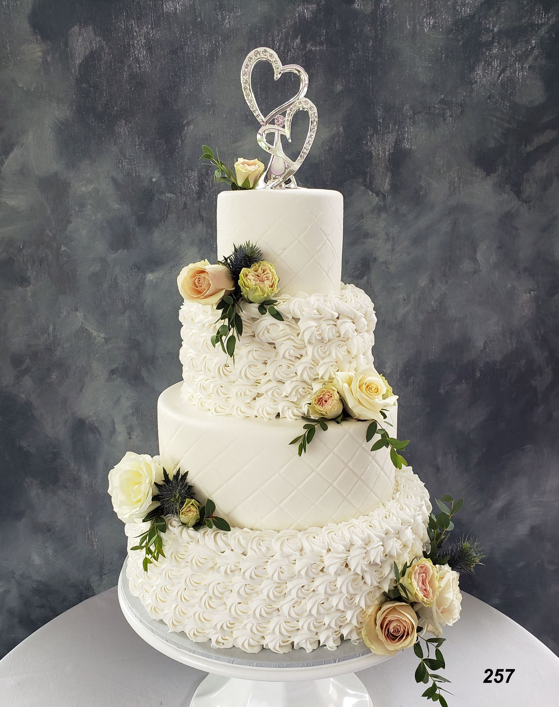Three-tier white wedding cake with floral decorations and heart toppers on a gray background
