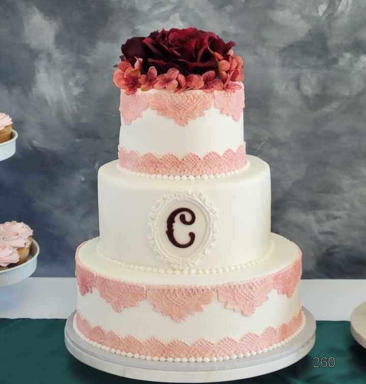 Three-tier white cake with pink lace trim and dark red flowers on top, featuring a monogrammed oval plaque