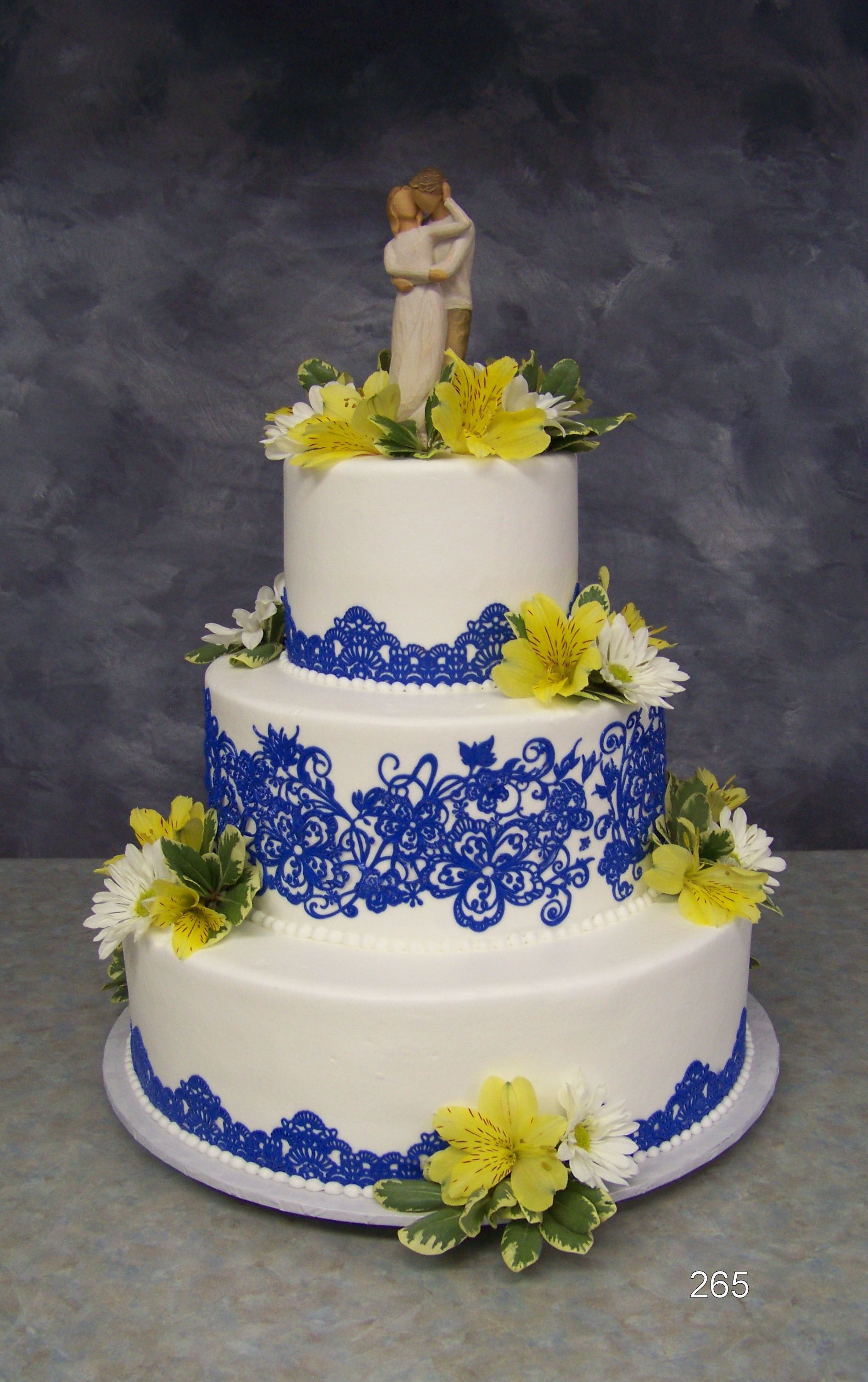 Three-tier white wedding cake with blue lace accents, yellow flowers, and a bride and groom topper