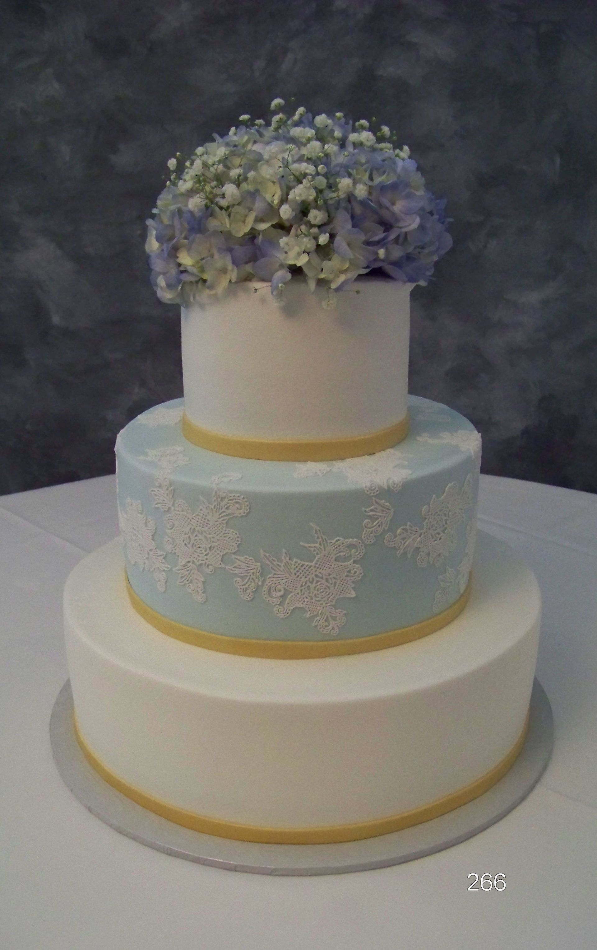 Three-tier white and pale blue wedding cake with yellow trim and a bouquet of blue and white flowers on top