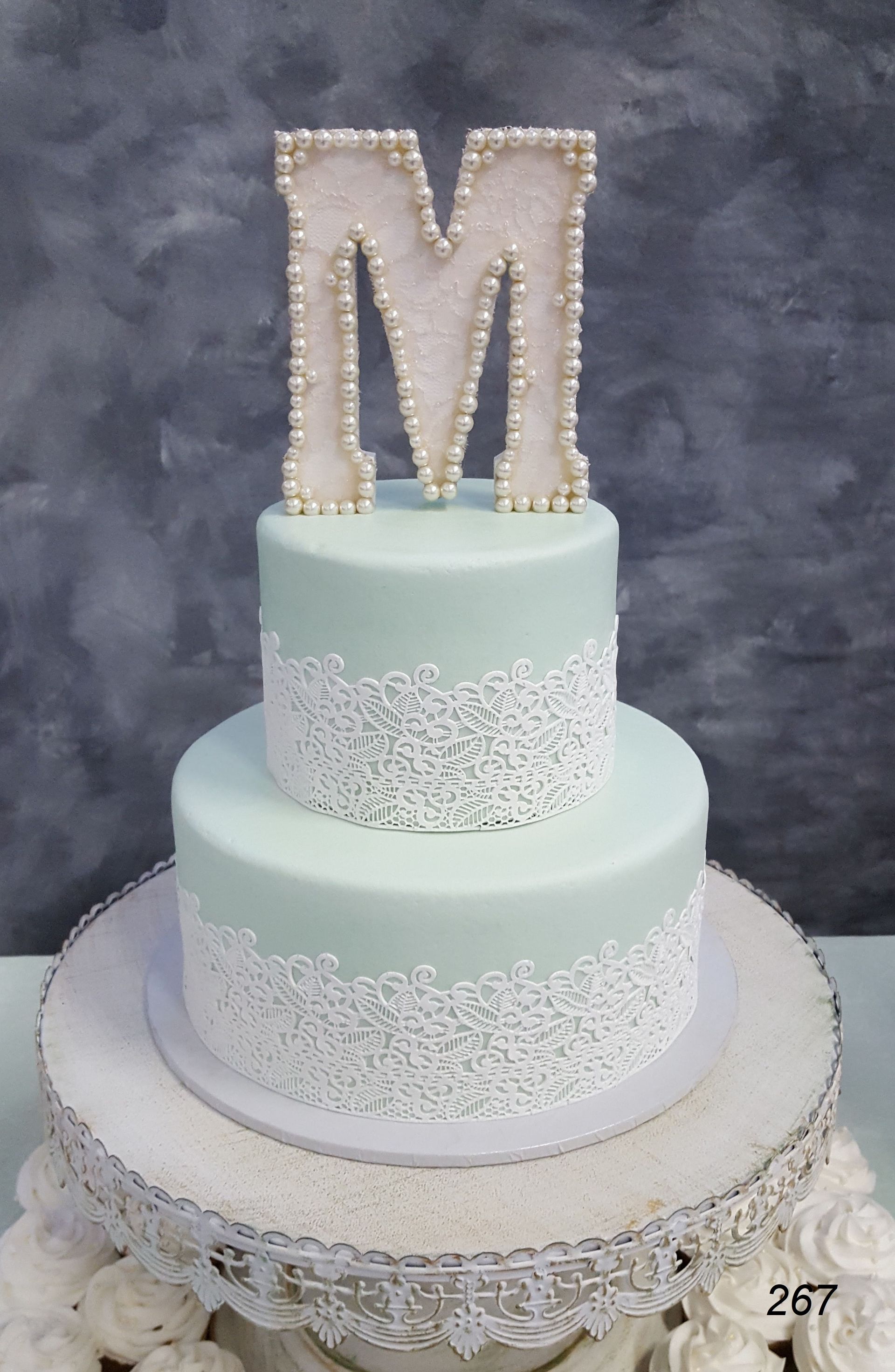 Three-tier white cake with pale blue accents and a jeweled letter M topper